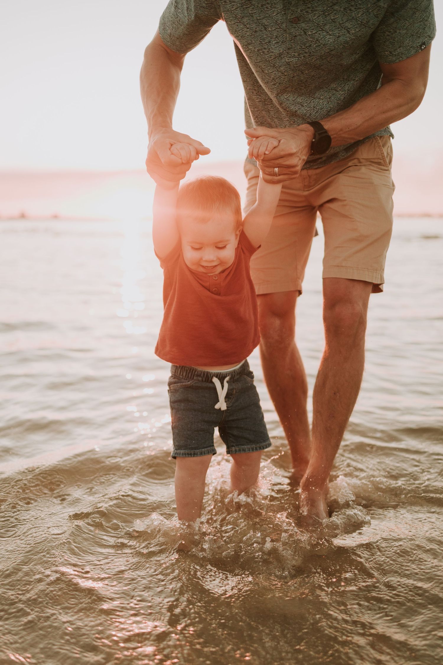 Father holding toddler's hands while walking in shallow ocean water at sunset, both wearing casual summer clothes.