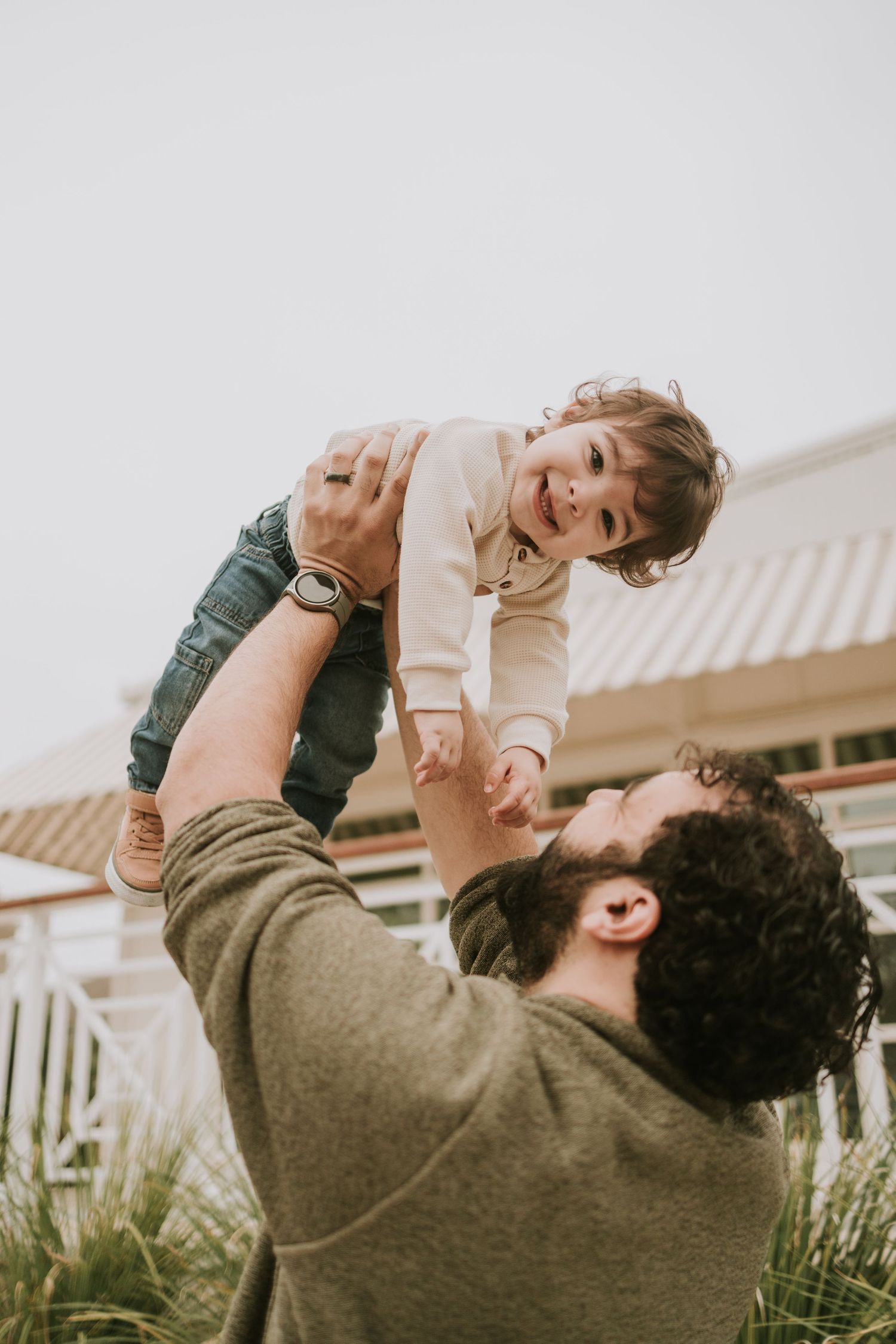 A bearded father lifts his smiling toddler high in the air outdoors, both sharing a joyful moment together.