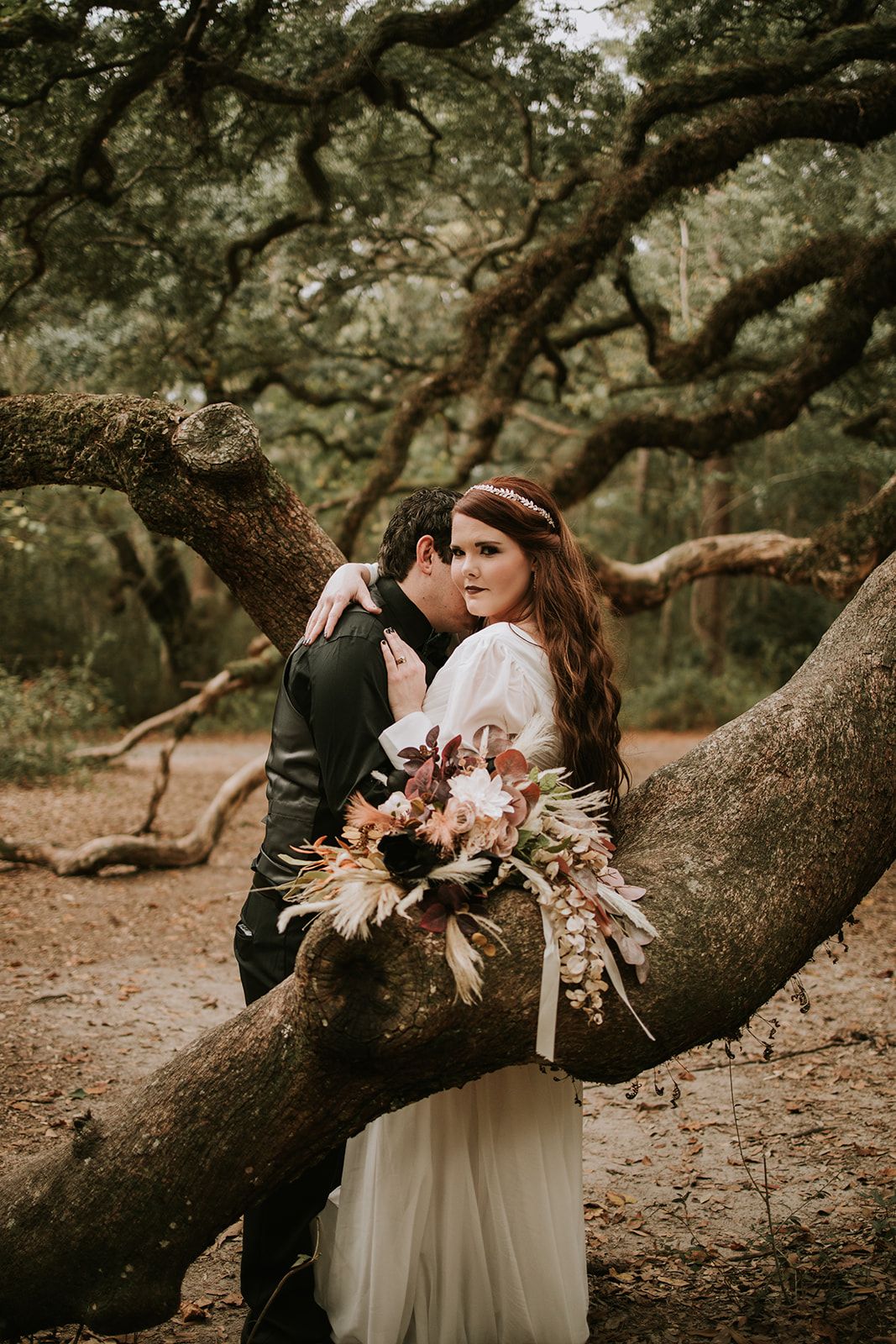 Romantic wedding portrait of couple embracing on large fallen tree branch with autumn-themed bouquet in forest setting.