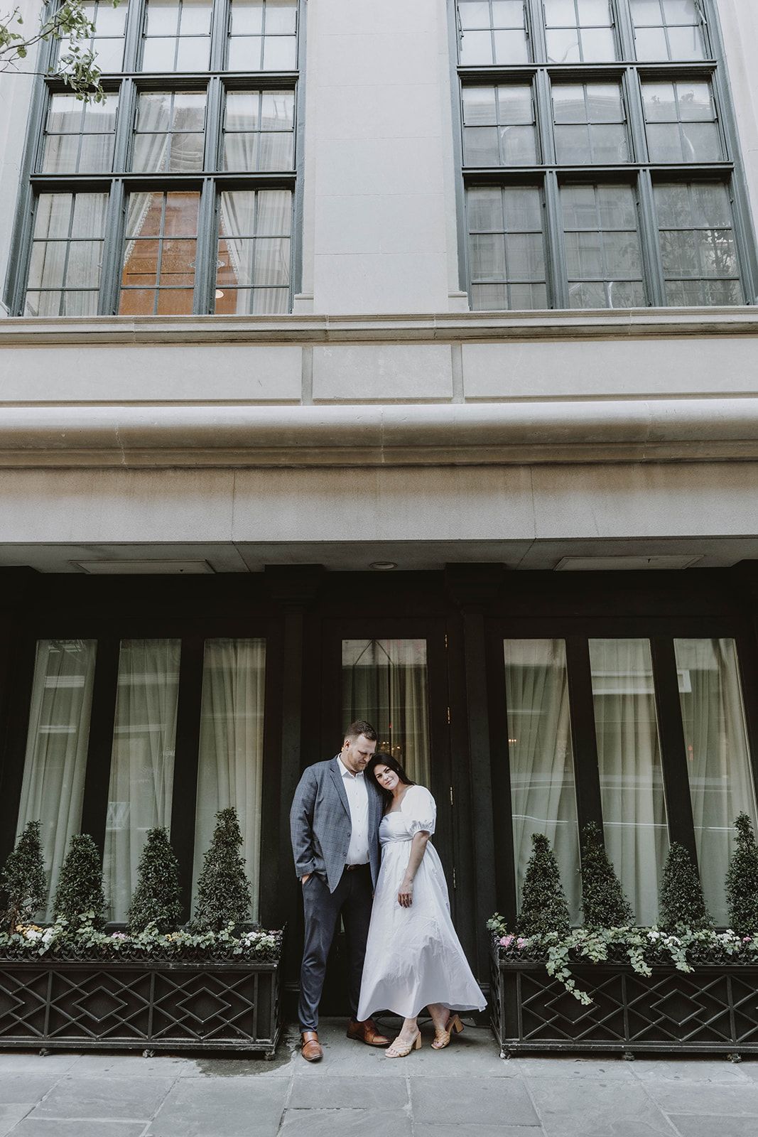 Elegant couple poses against urban building exterior with large windows and decorative architecture in vintage style setting.