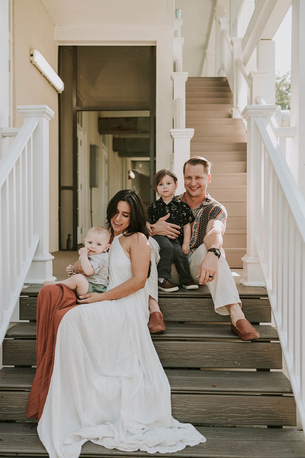 Family portrait on rustic white porch steps with parents and two small children in light-colored clothing.