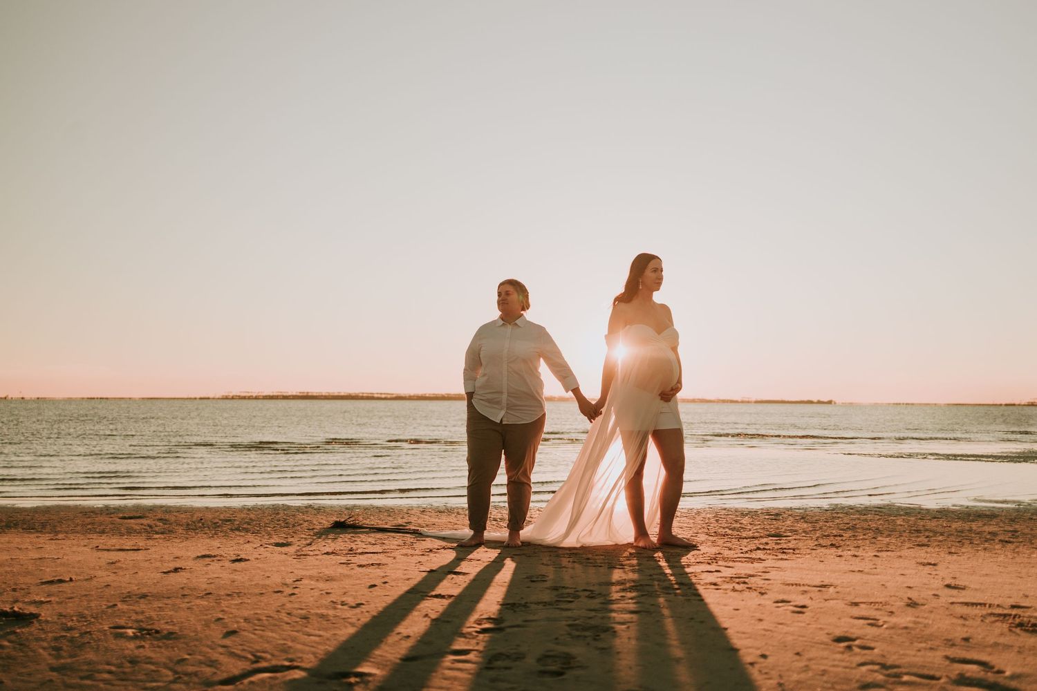 A couple holding hands and casting long shadows on a beach during a golden sunset over the ocean.