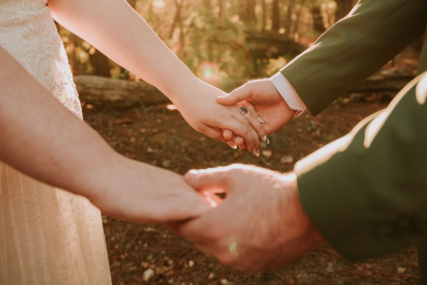 Two people hold hands intimately during a romantic golden hour moment in a wooded setting.