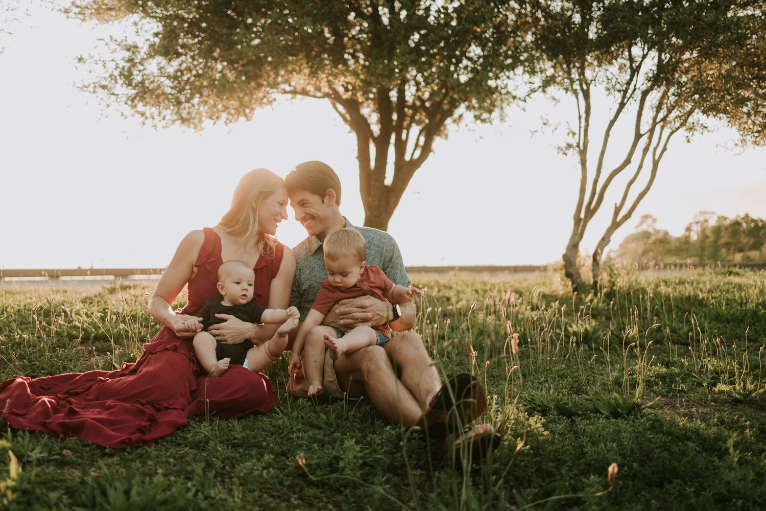 Family of four sitting together on lush green grass at golden hour beneath trees, parents gazing lovingly at two young children.
