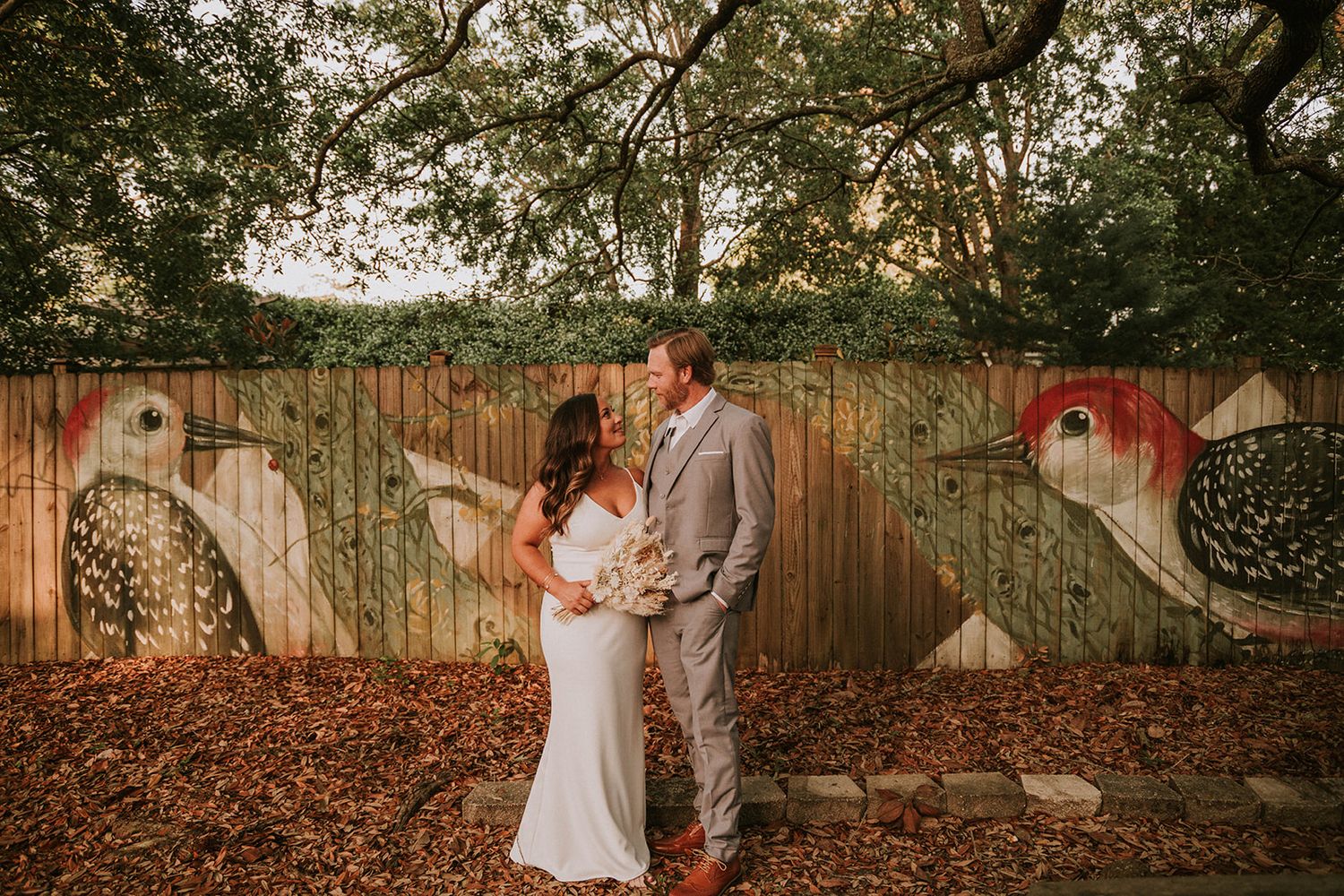 A romantic outdoor wedding moment by a wooden fence decorated with large bird murals in a garden setting at sunset.