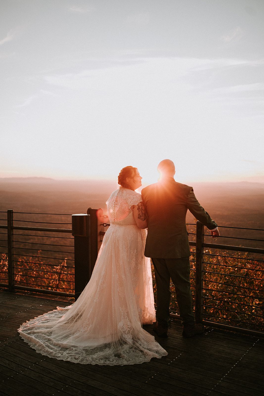 A couple in wedding attire stands on a deck overlooking mountains at sunset, their silhouettes backlit by golden light.