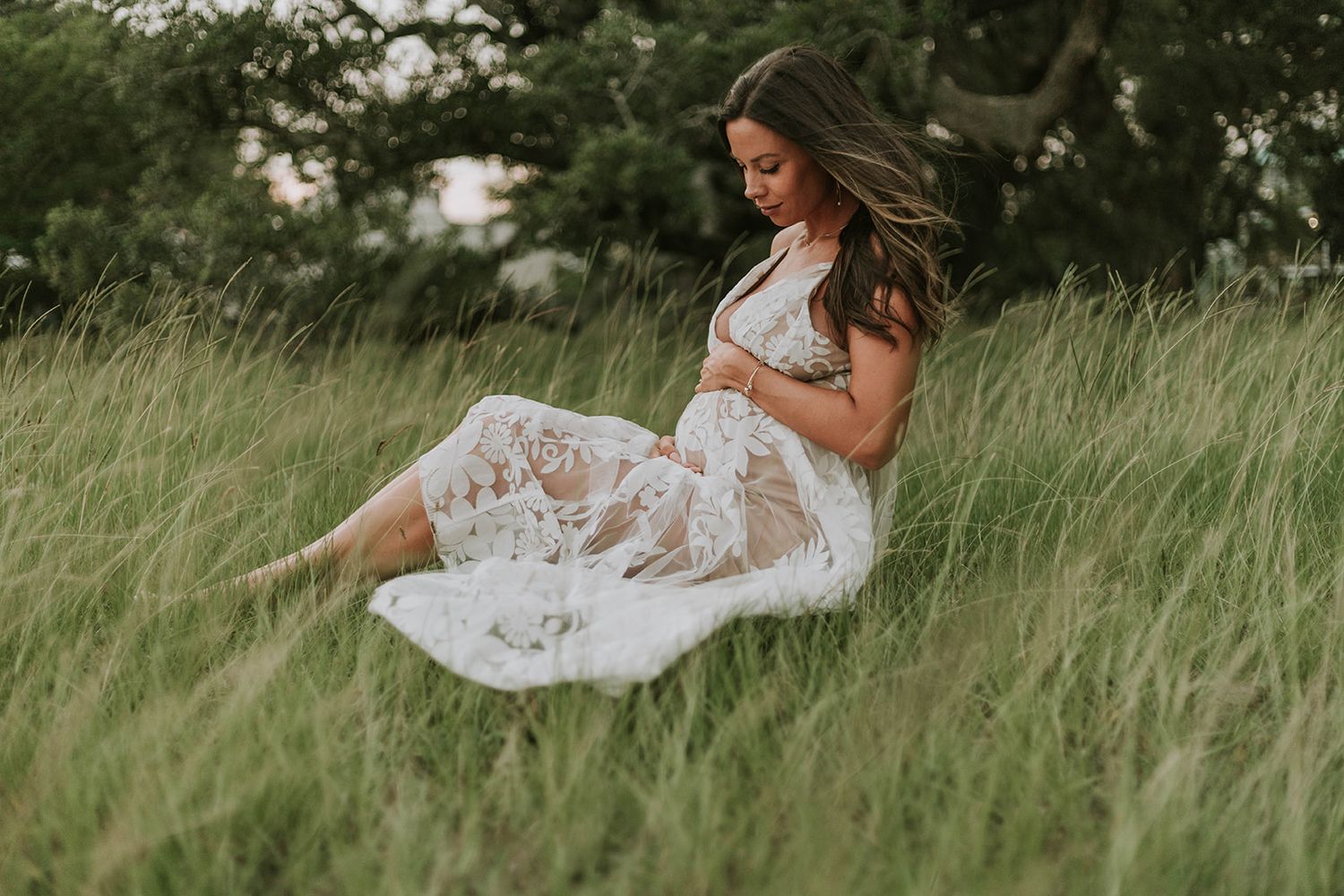 Ethereal maternity portrait of woman in white lace dress sitting in meadow.