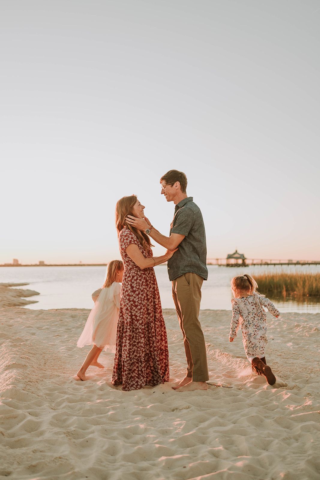 A family shares a tender moment together on a sandy beach at sunset with marsh grass in the background.