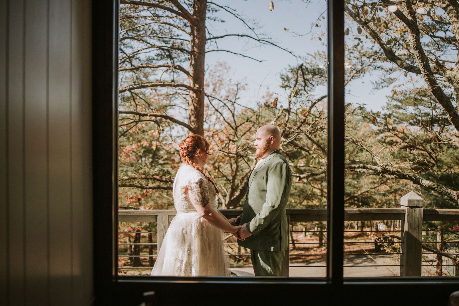 A couple shares an intimate outdoor moment on a deck surrounded by autumn trees, viewed through a window at sunset.