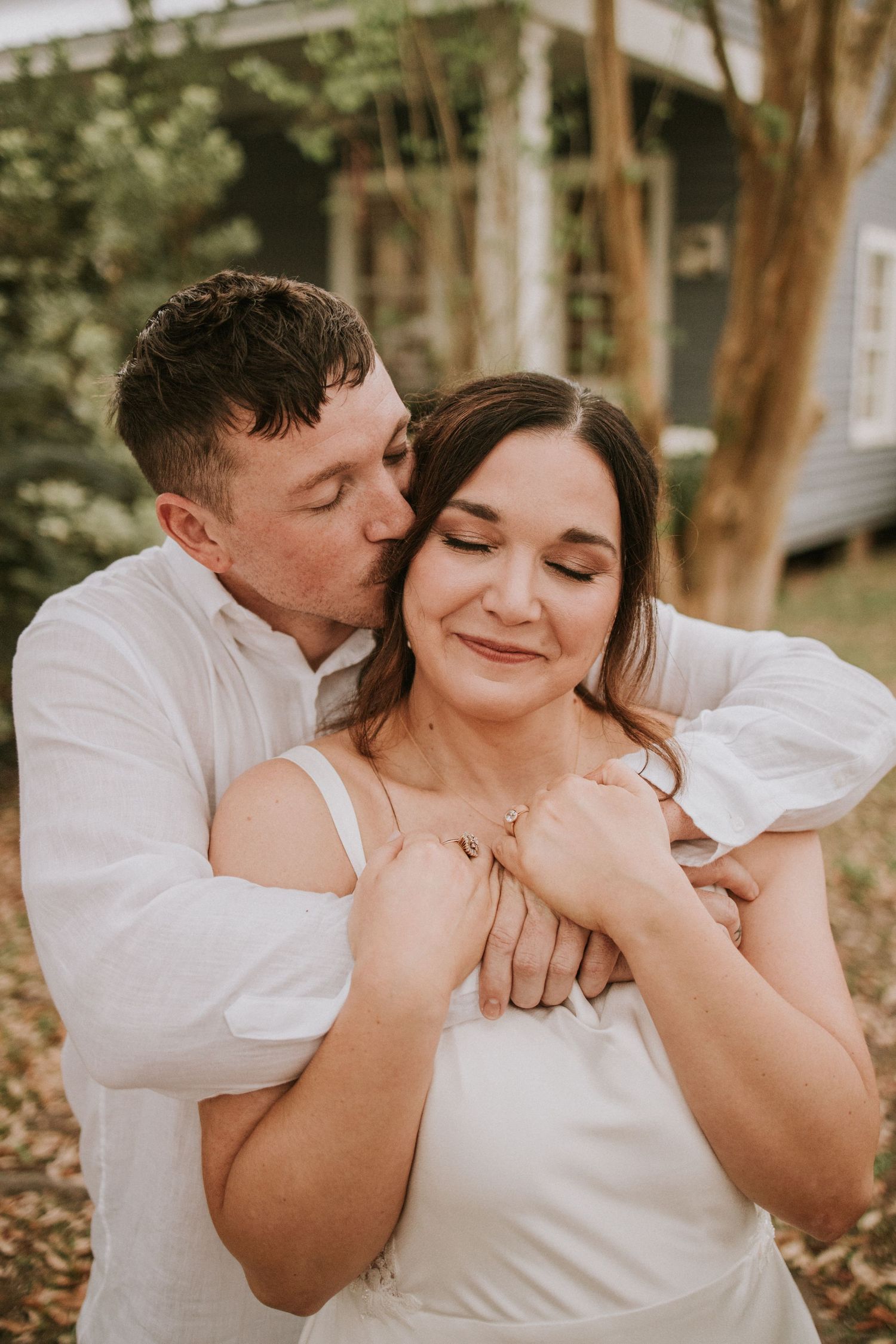 A couple in white embraces outdoors, man kissing woman's cheek while holding her hands near a tree.