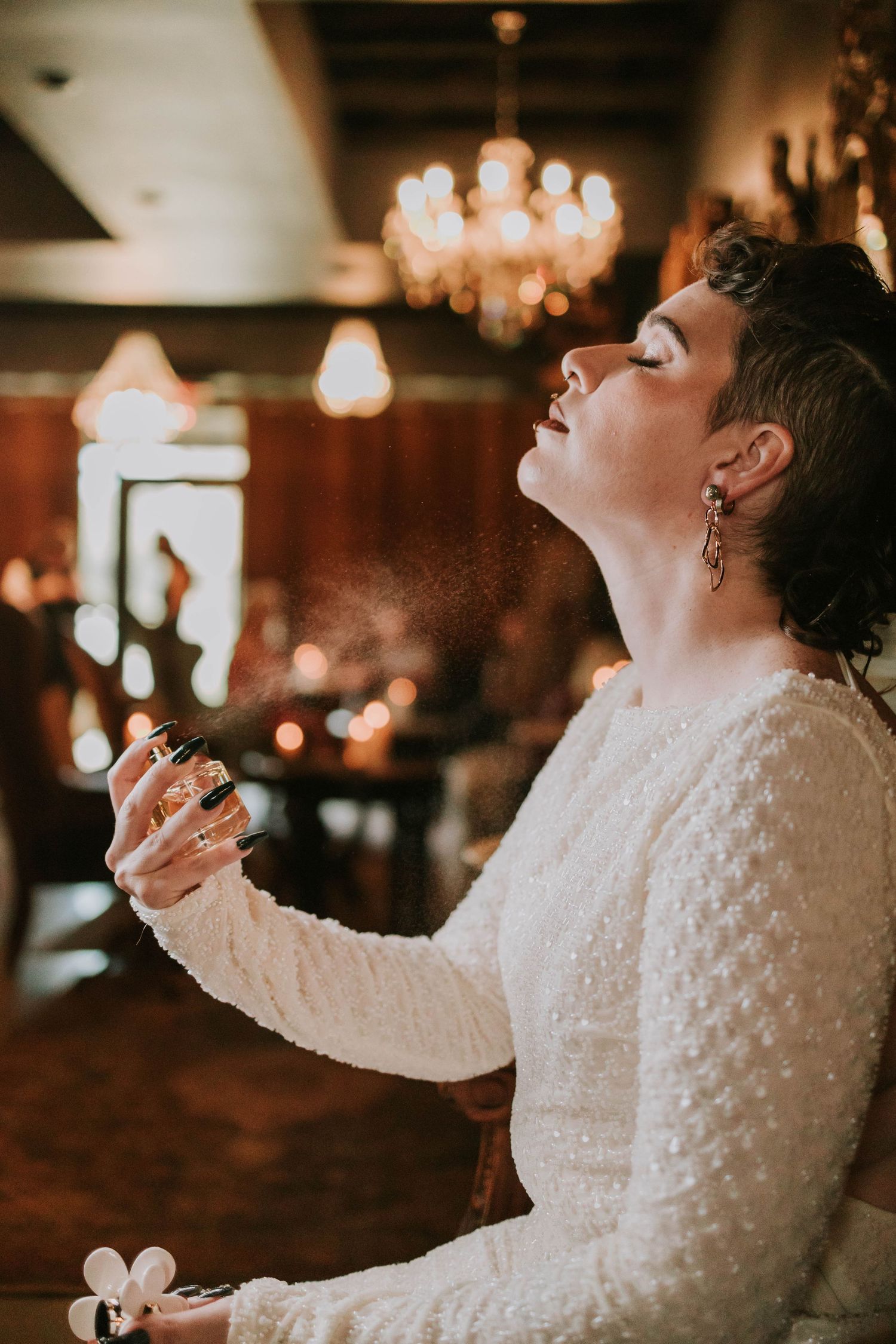 Bride in white lace dress spraying perfume with eyes closed in elegant chandelier-lit venue.