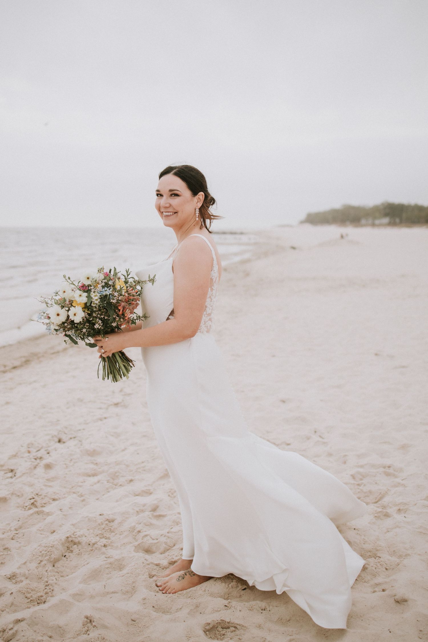 Smiling bride in white gown holding colorful bouquet on sandy beach with calm ocean in background.