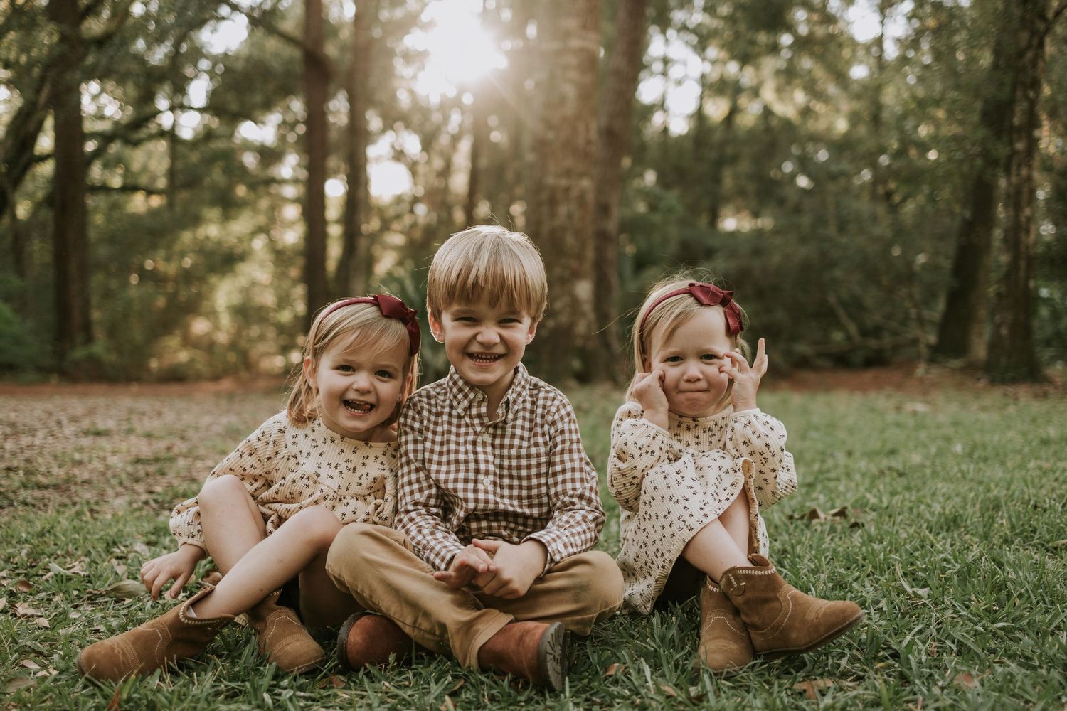 Three young siblings smile and sit together on green grass in a sunlit outdoor park setting.