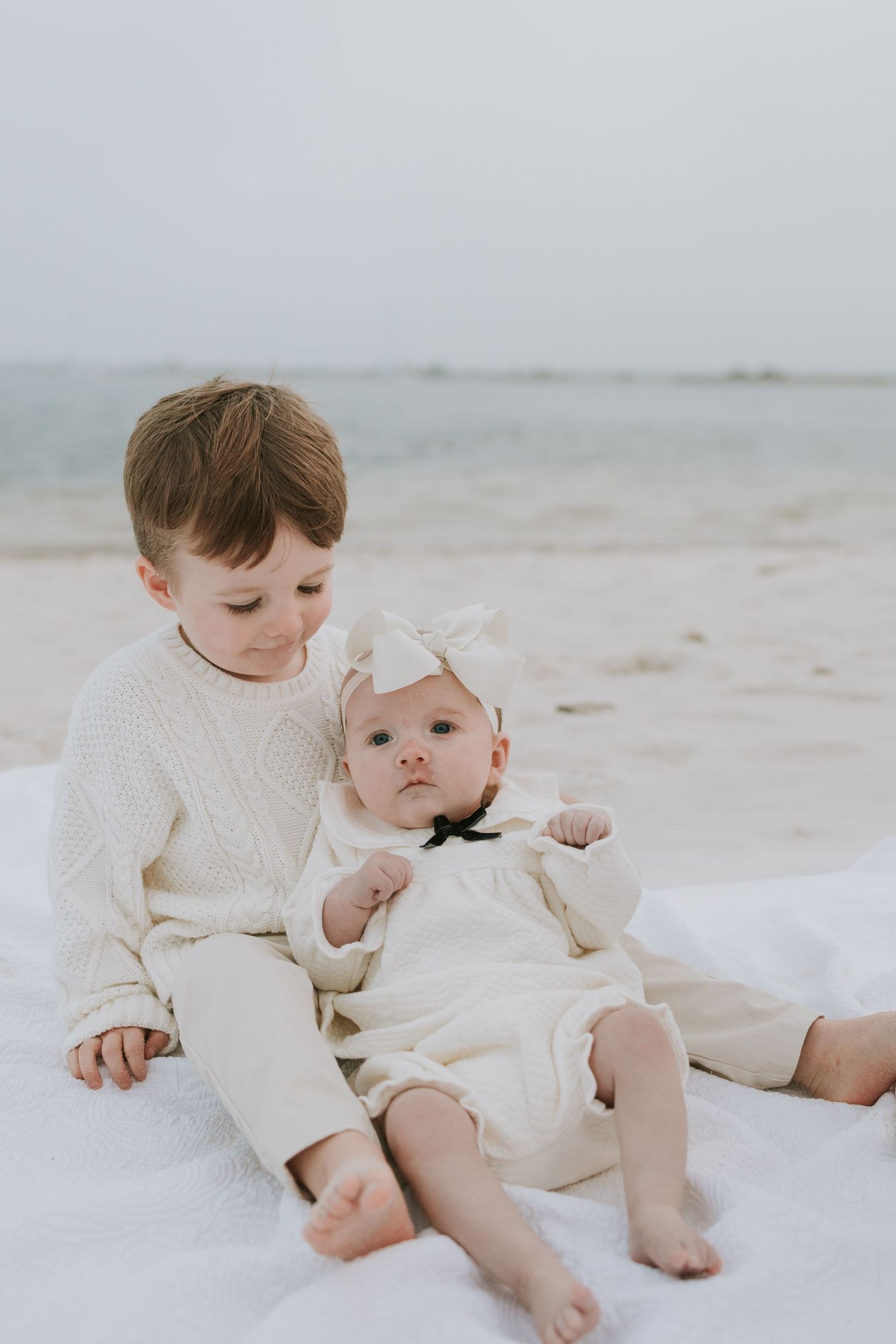 Two young siblings in cream outfits sit on a white blanket at the beach, older boy gently holding baby girl.
