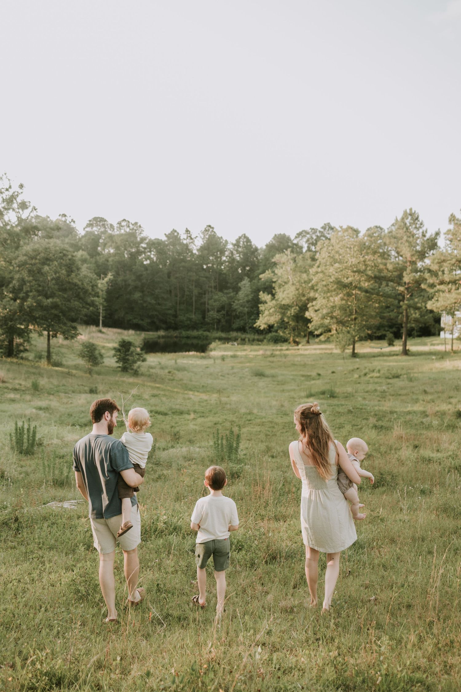 A family of five walks through a lush green meadow surrounded by trees, with parents carrying young children.