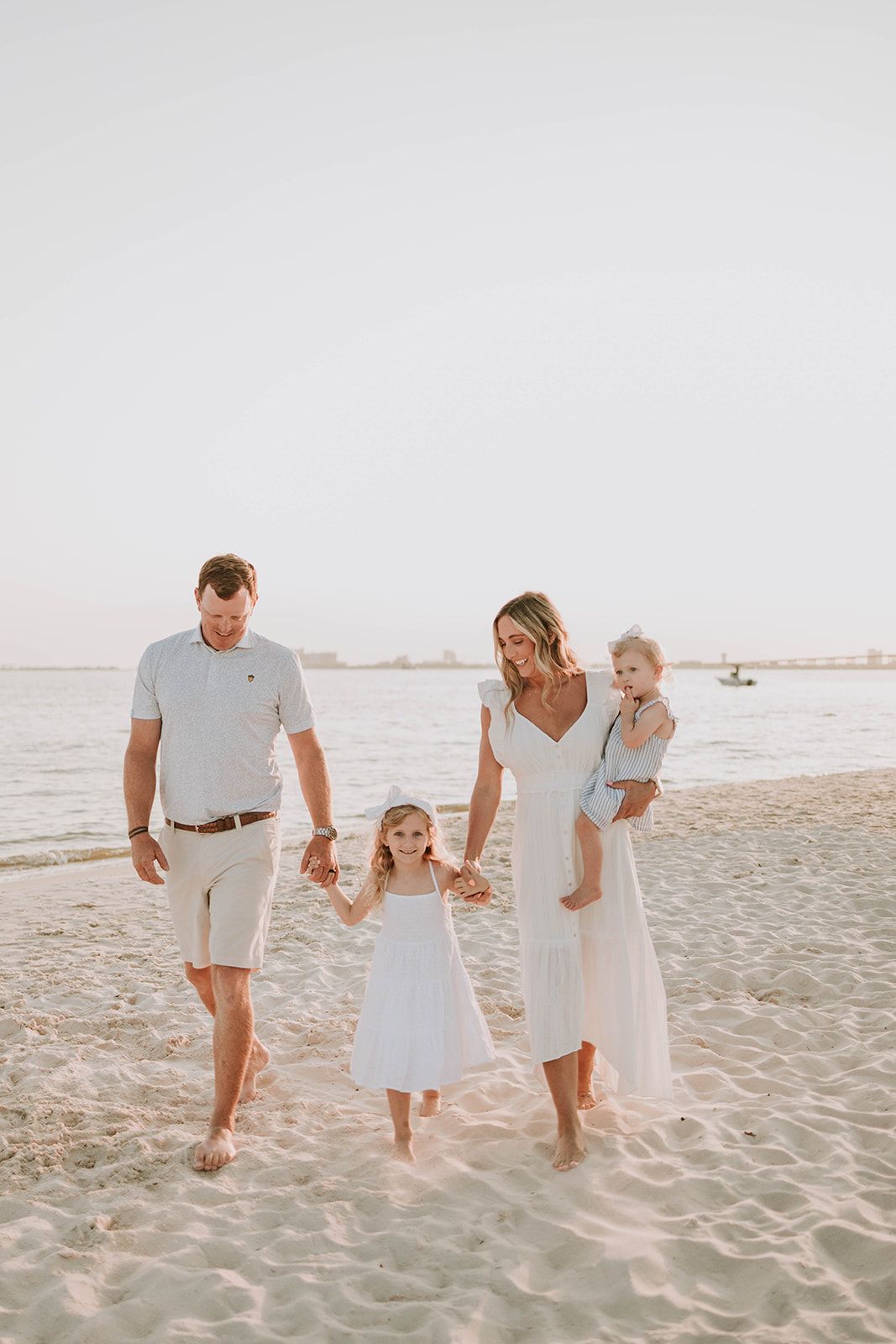 Family wearing light colored outfits walks together on a beach at sunset with water and boats visible in background.