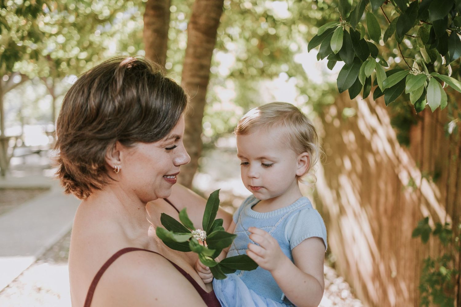 In a sunlit garden setting, a caregiver and young child examine green leaves together while sharing a tender moment.