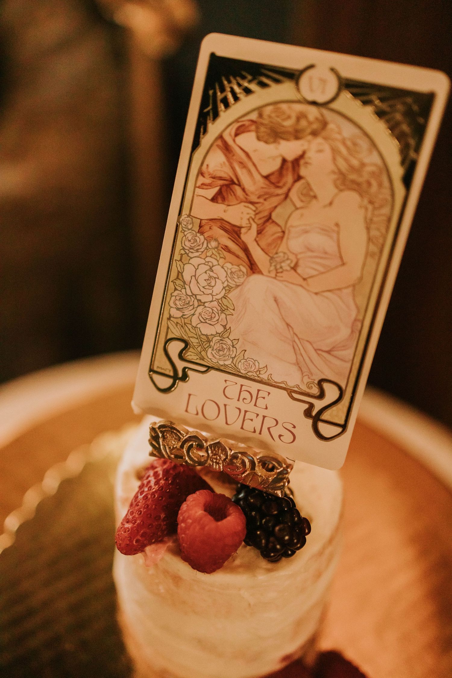 A tarot card 'The Lovers' displayed on a small white cake topped with fresh berries in warm ambient lighting.