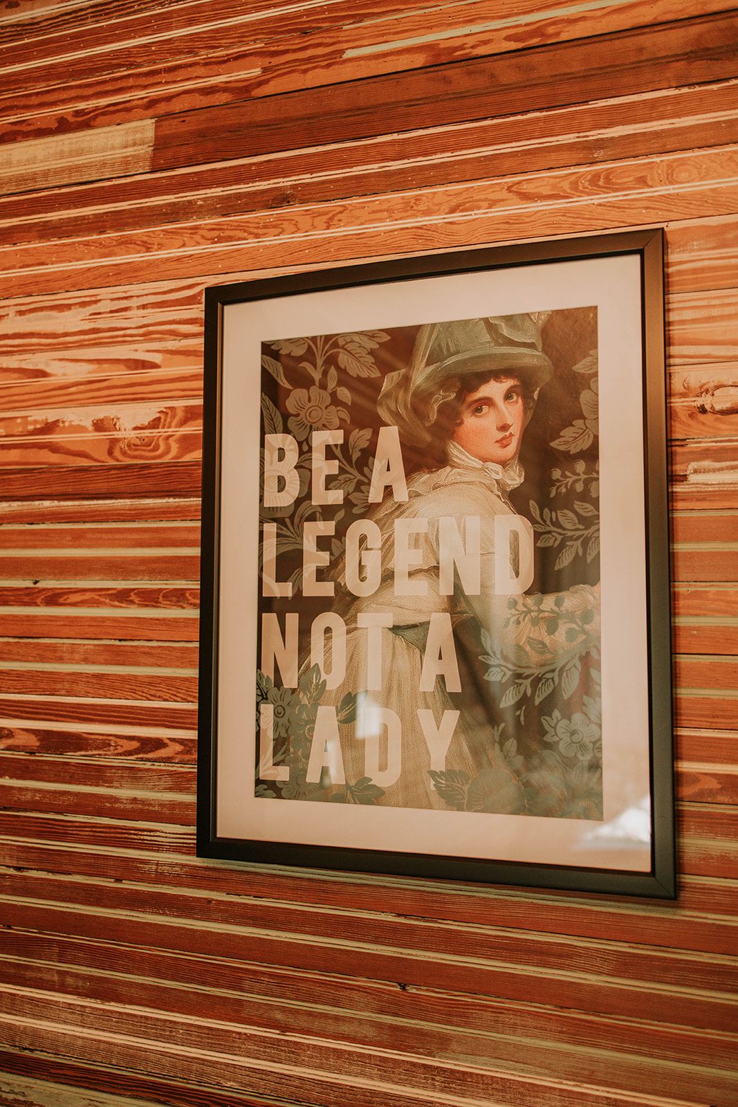 A framed vintage-style artwork displaying 'BE A LEGEND NOT A LADY' text hangs on a wooden paneled wall.