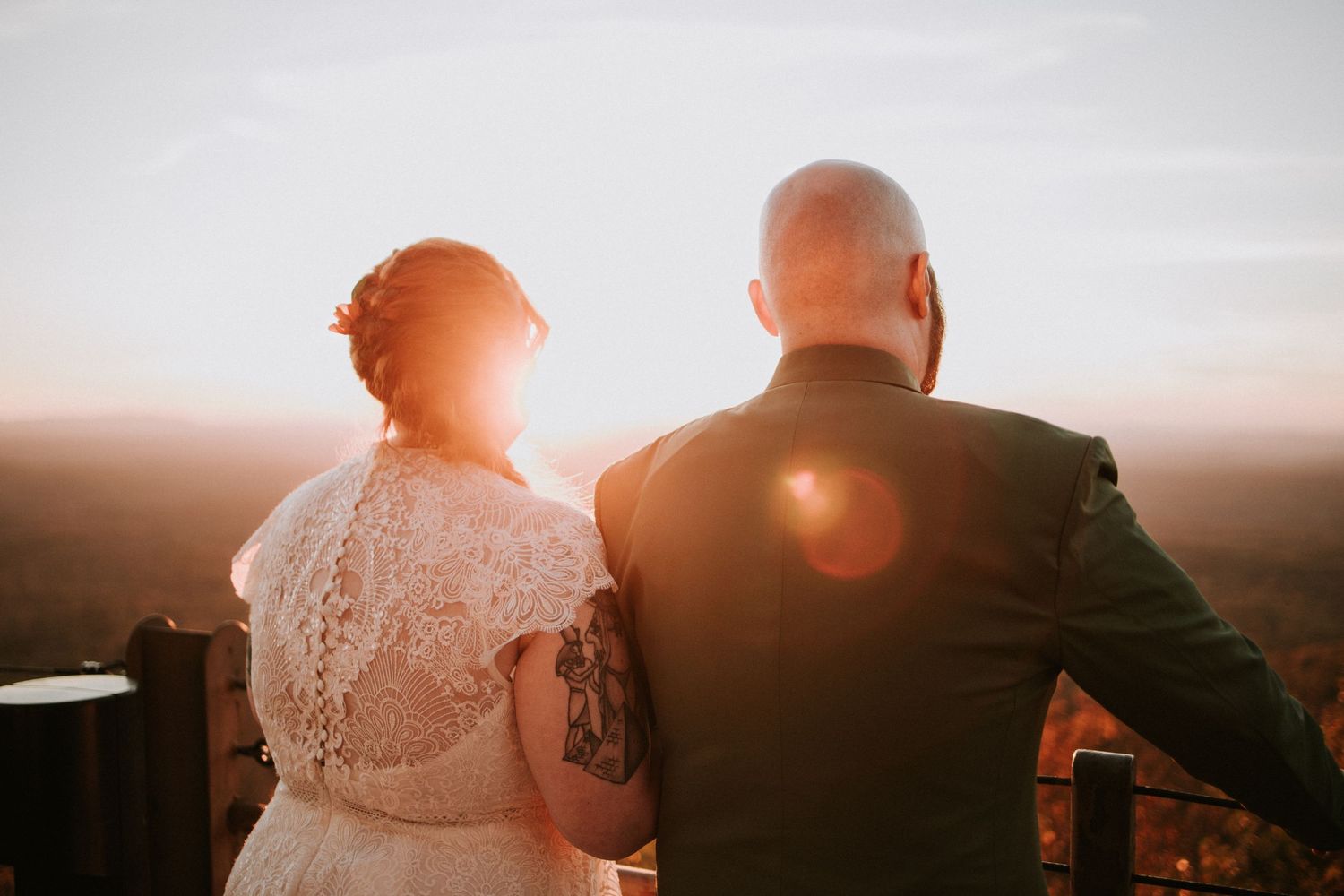 A couple sits together watching the sunset, with sunflare visible as the bride wears a lace dress and groom in dark suit.