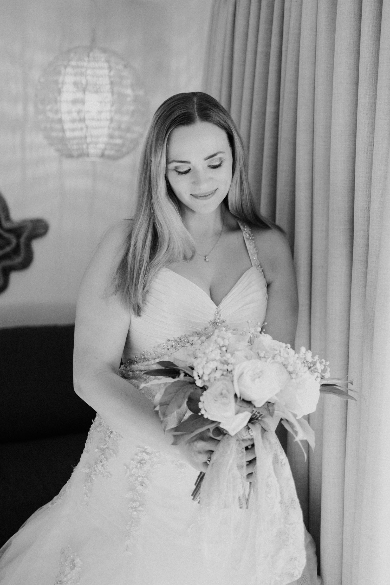 Bride in strapless lace wedding gown holds floral bouquet, smiling softly in black and white portrait photo.