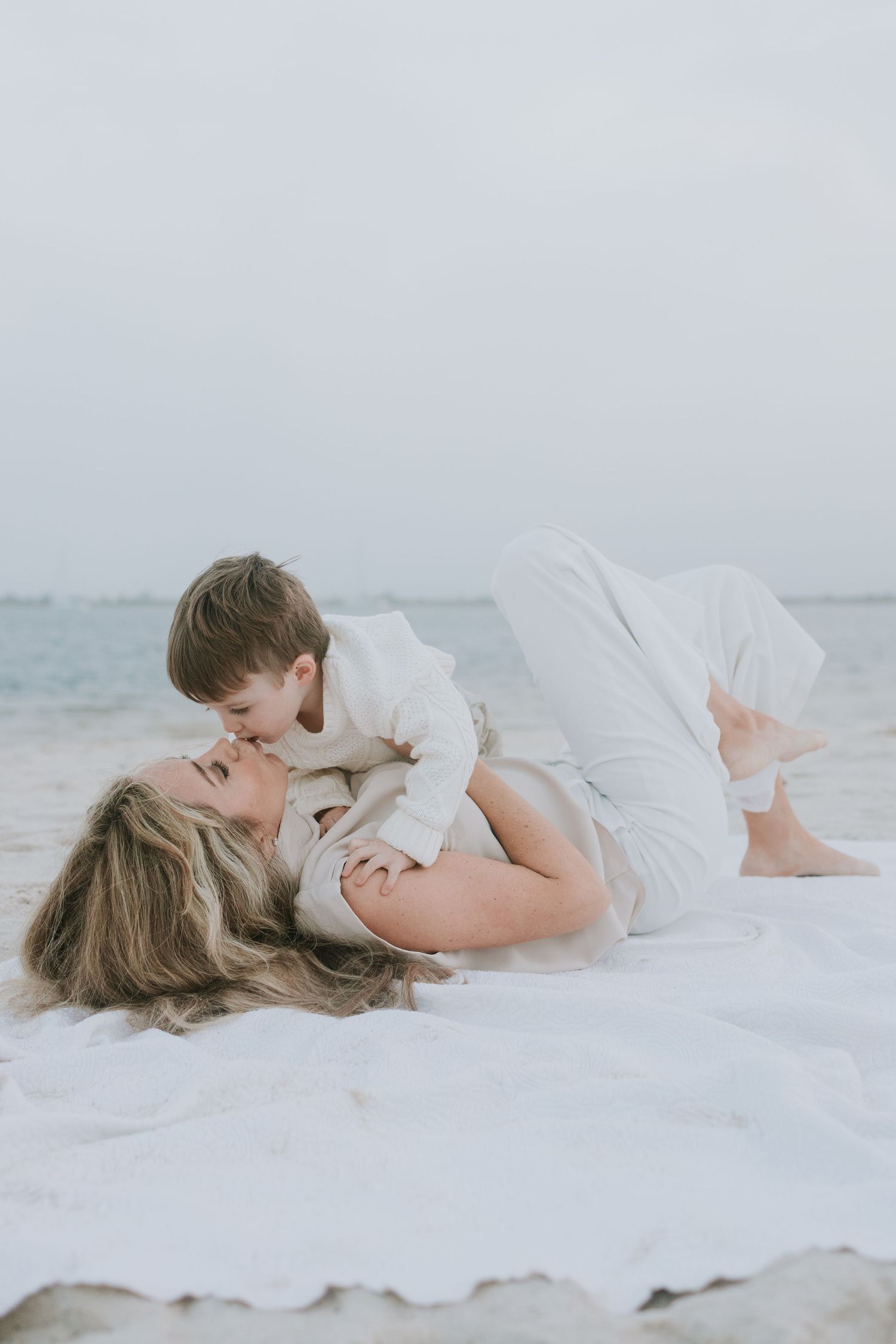 Mother lying on beach blanket kissing young child in matching white outfits with ocean in background.