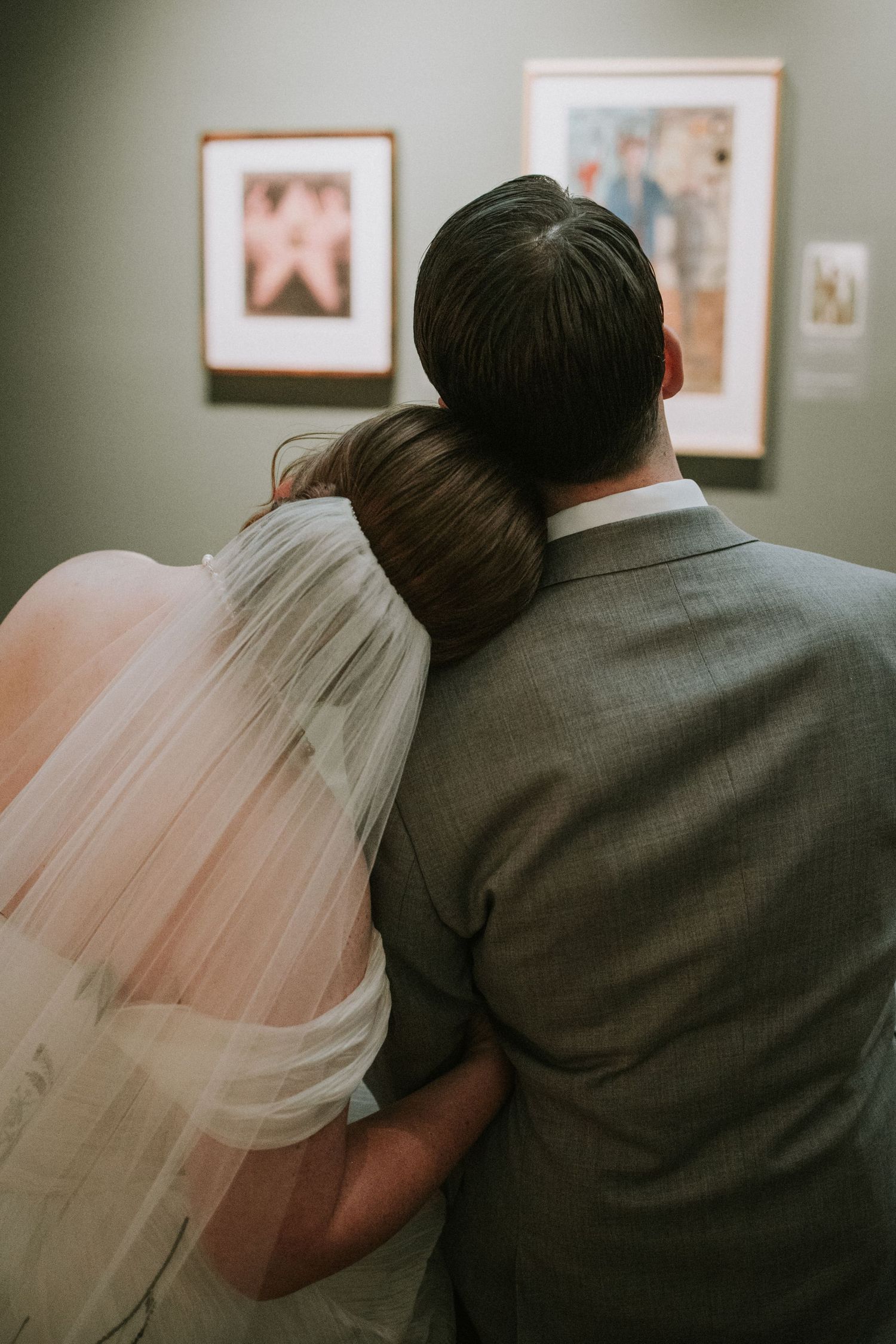 Bride and groom viewed from behind, embracing while admiring framed artwork on gallery walls at their wedding.