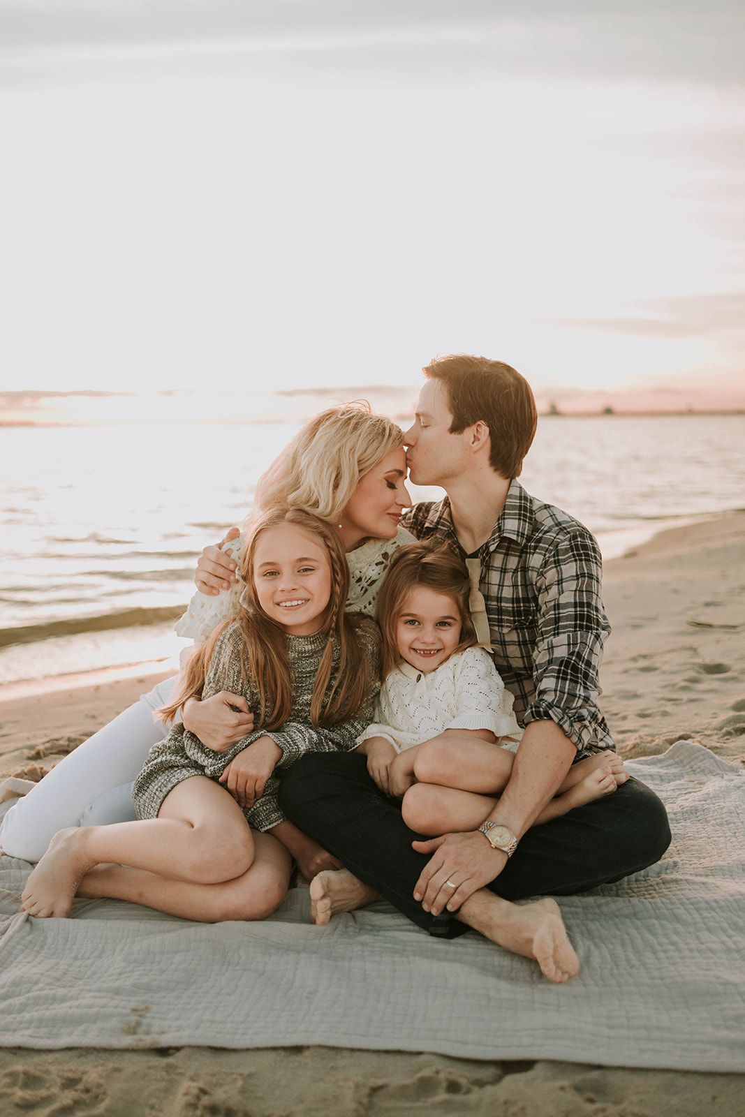 Family cuddling together on blanket at beach during sunset.