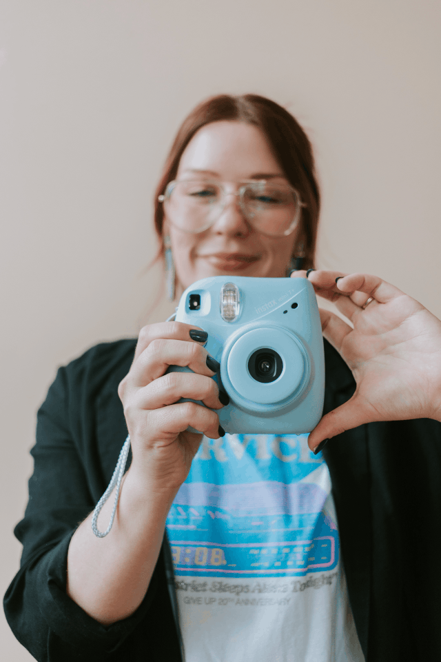 Woman with glasses holds a light blue Fujifilm Instax Mini camera up to take a selfie in a bright room.