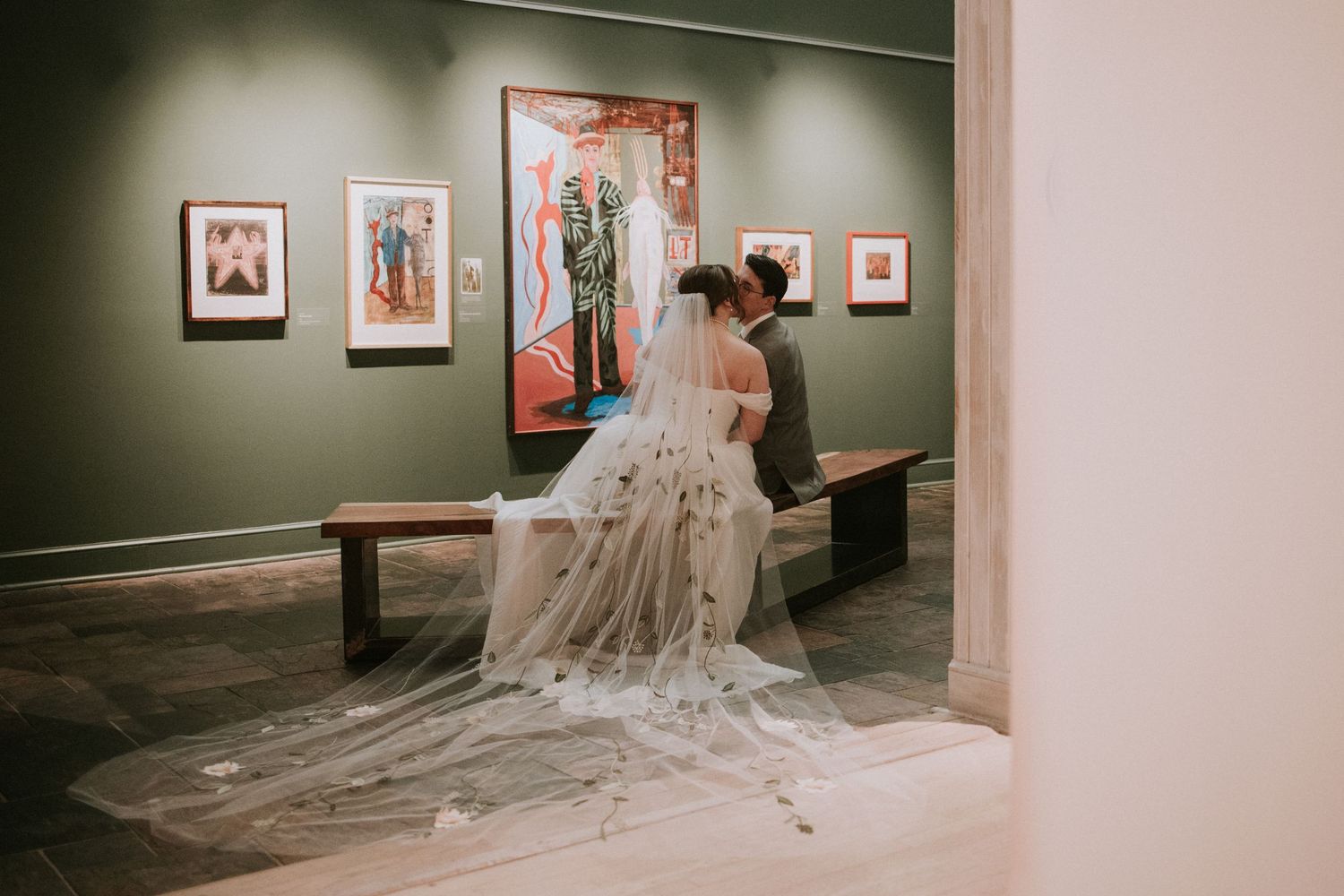 Bride and groom share an intimate moment in an art gallery, her long lace veil trailing elegantly on the floor.