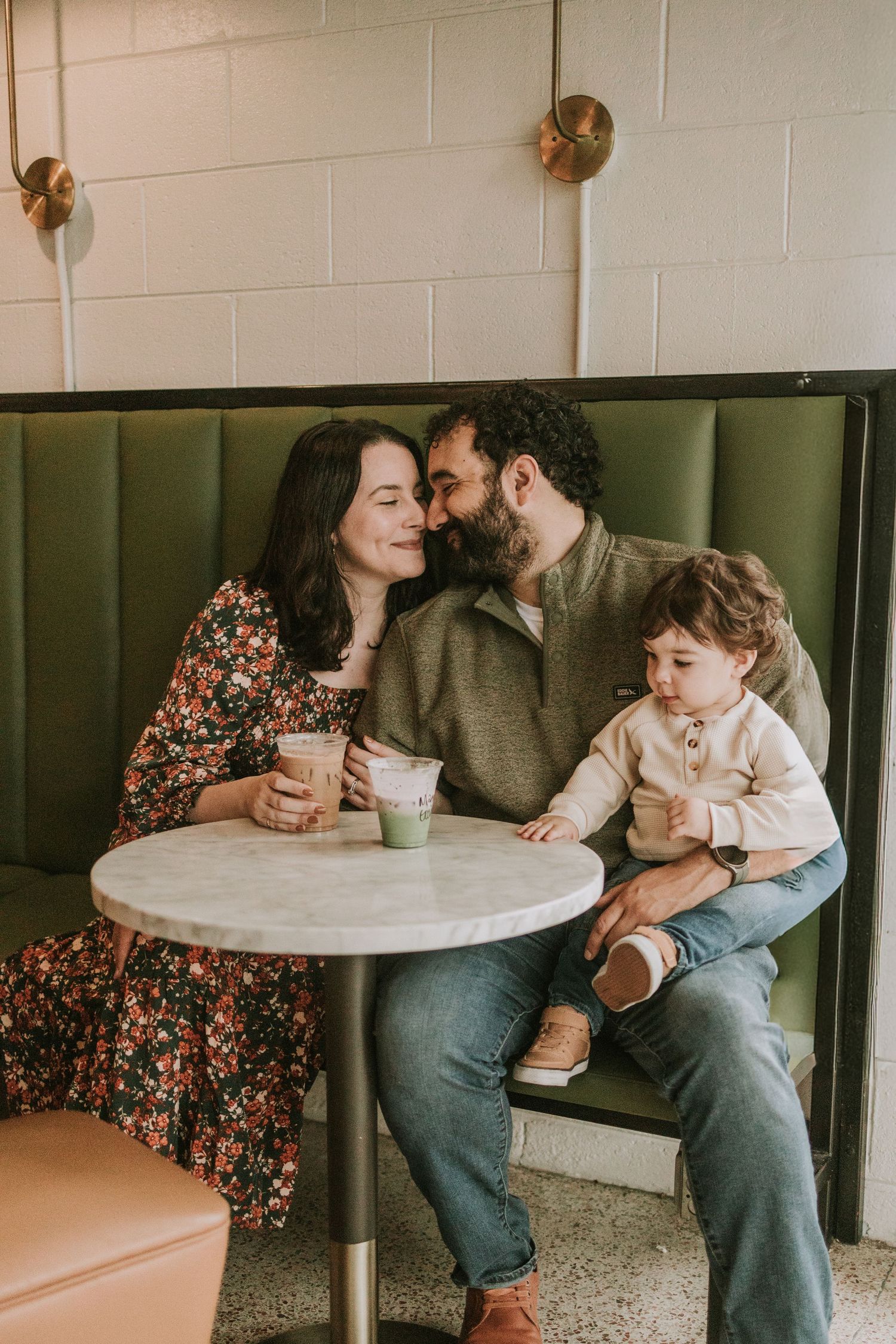 A loving family sits in a cozy cafe booth, parents sharing a kiss while holding a toddler and green drinks.
