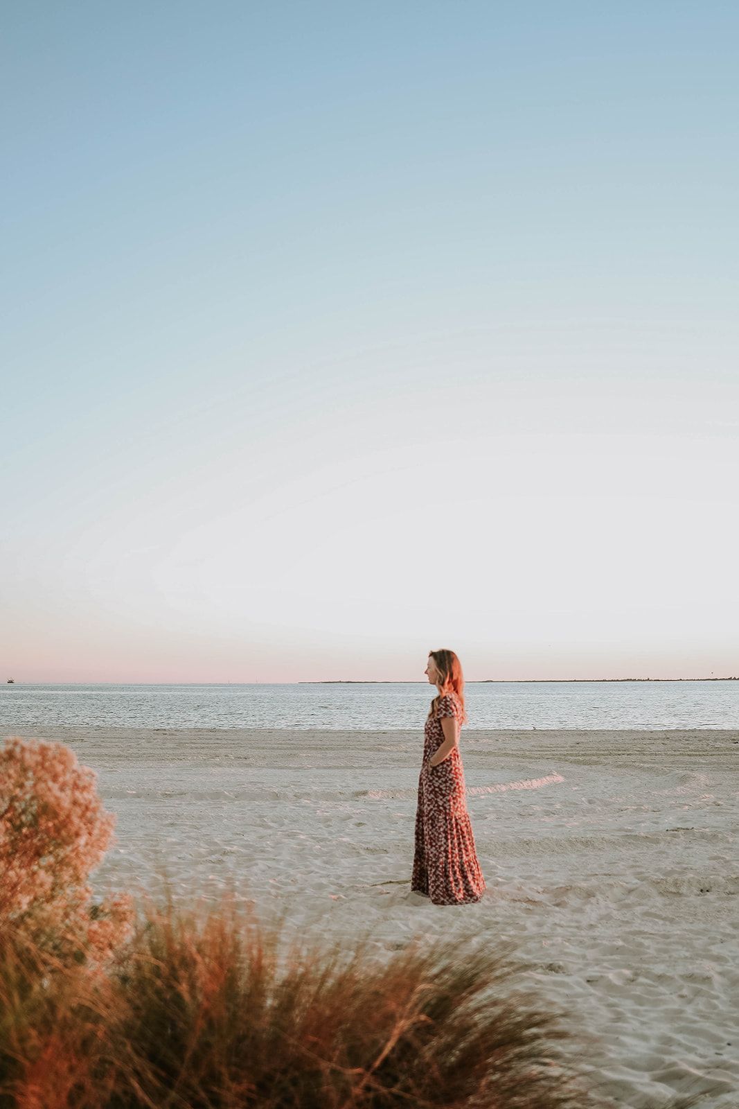 A lone woman in a floral maxi dress stands on an empty beach during golden hour.