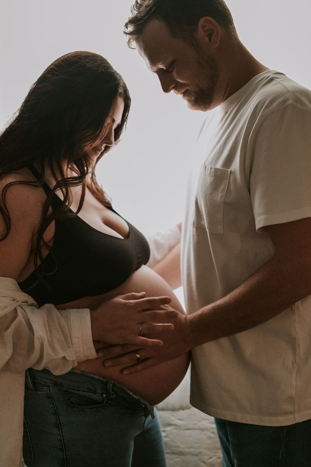 A couple shares an intimate moment looking down at her pregnant belly in a backlit maternity shoot.