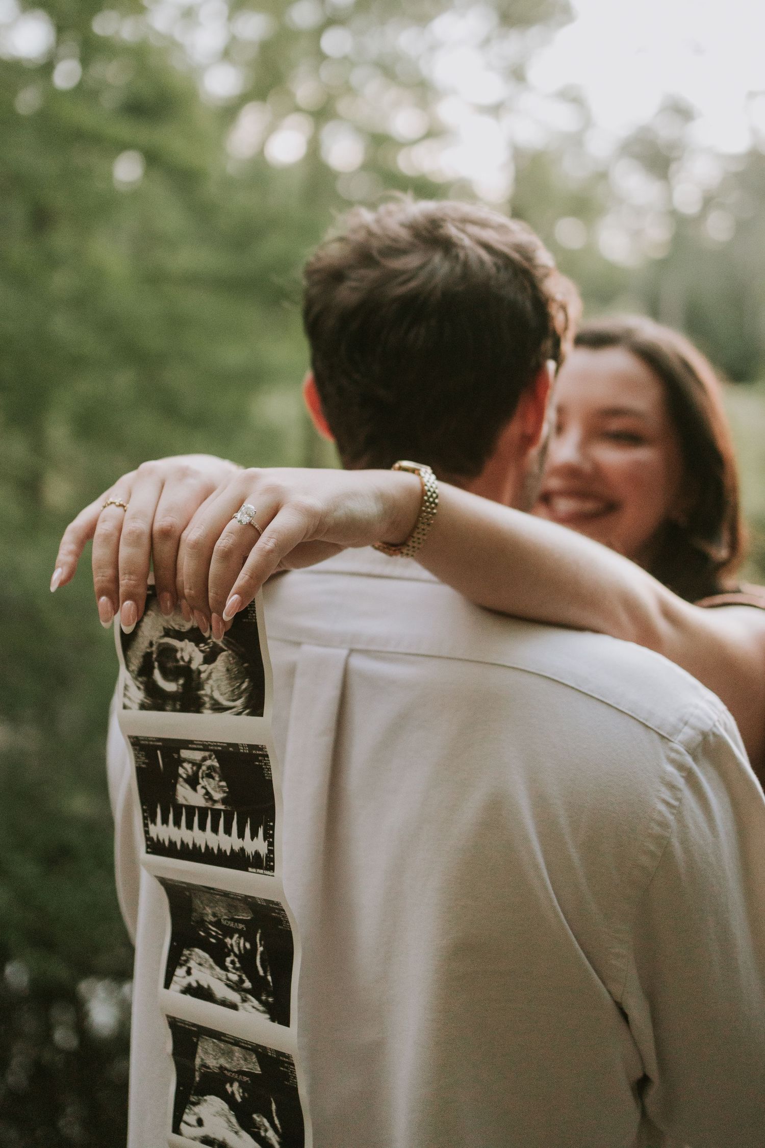 Couple embracing outdoors with ultrasound photos tucked in back pocket, announcing pregnancy with smiling woman visible.