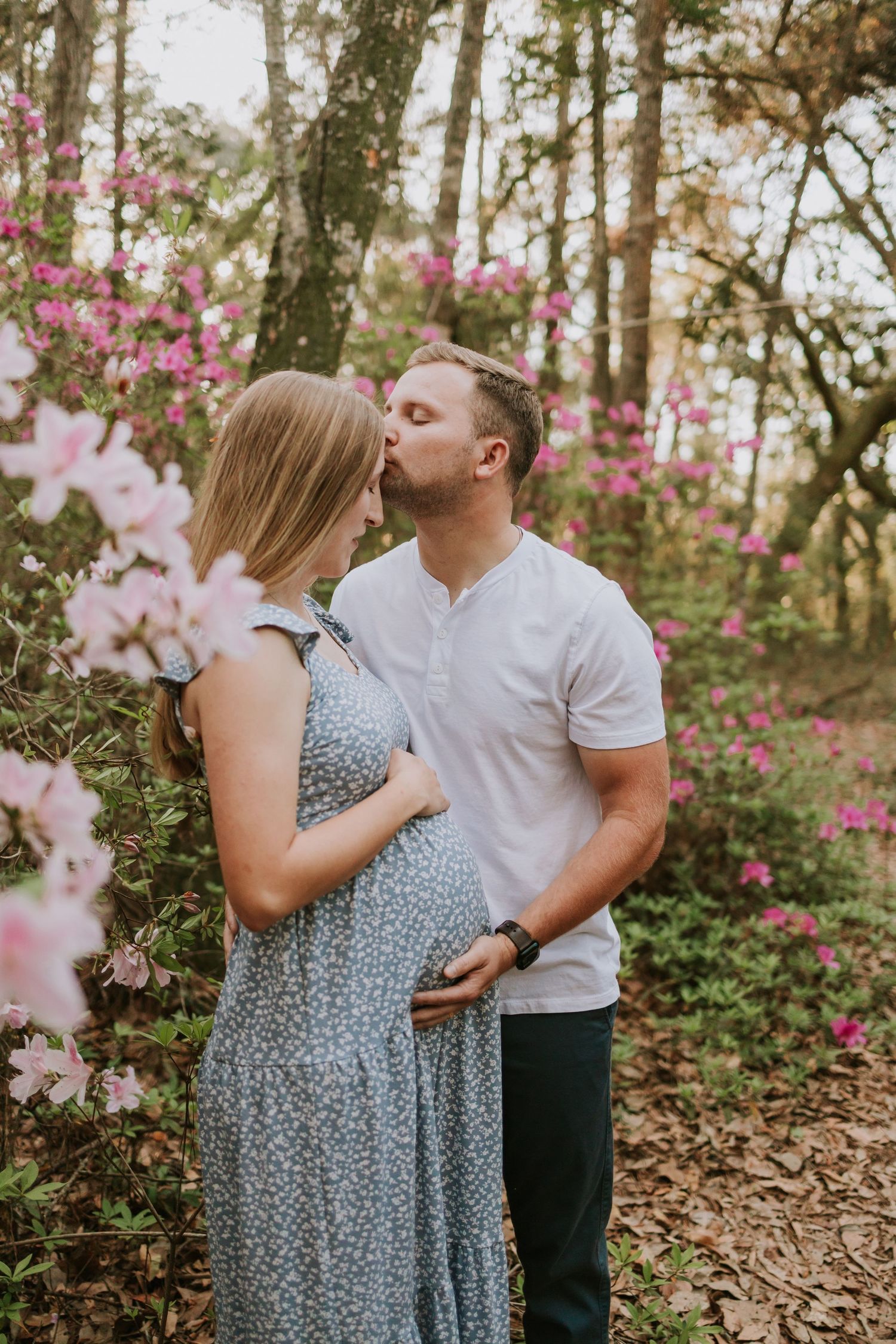 A couple shares a tender kiss outdoors among pink azalea blooms, with the woman in a blue floral dress cradling her pregnant belly.