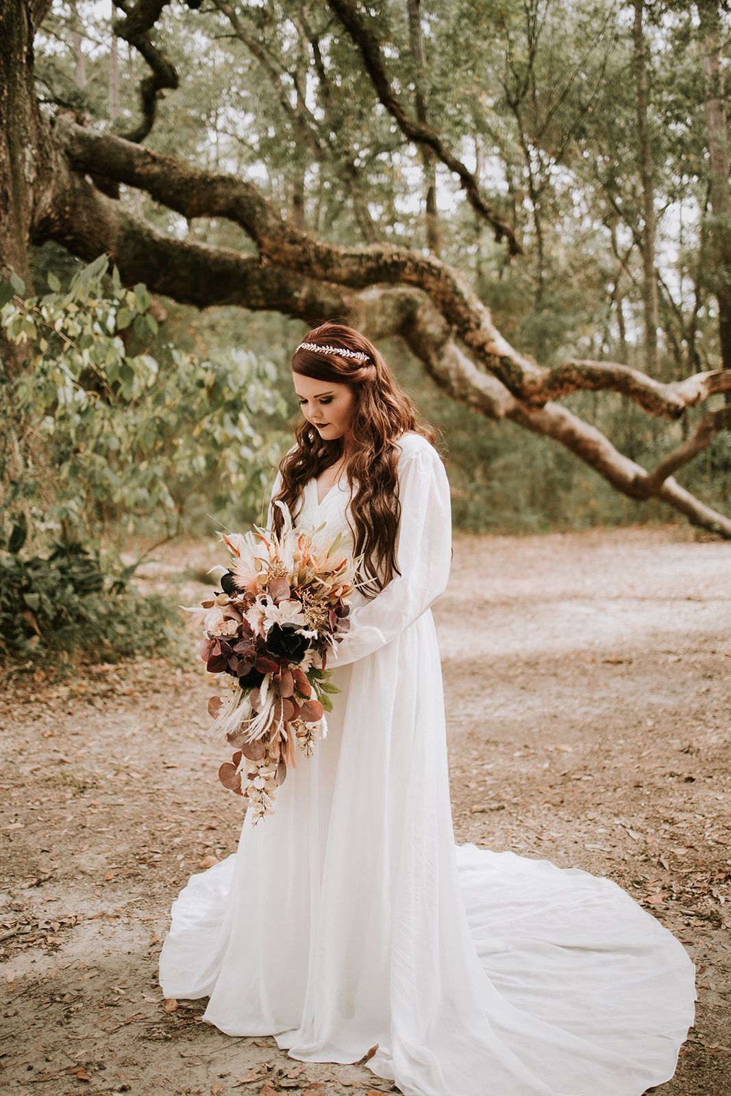A bridal portrait in a whimsical forest setting showing a long white dress and autumn-toned bouquet under twisted tree branches.