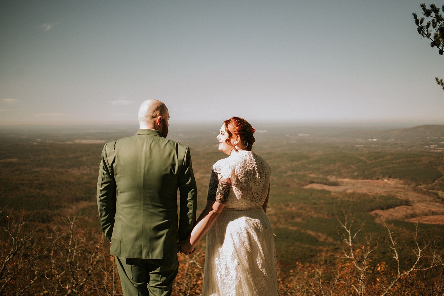 A newlywed couple stands together on a hilltop overlooking a vast landscape during golden hour.
