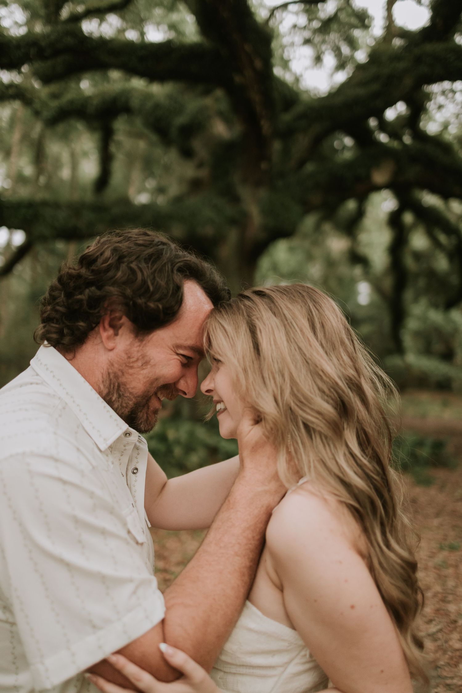A couple shares a romantic forehead-to-forehead moment, smiling tenderly, surrounded by lush green trees outdoors.