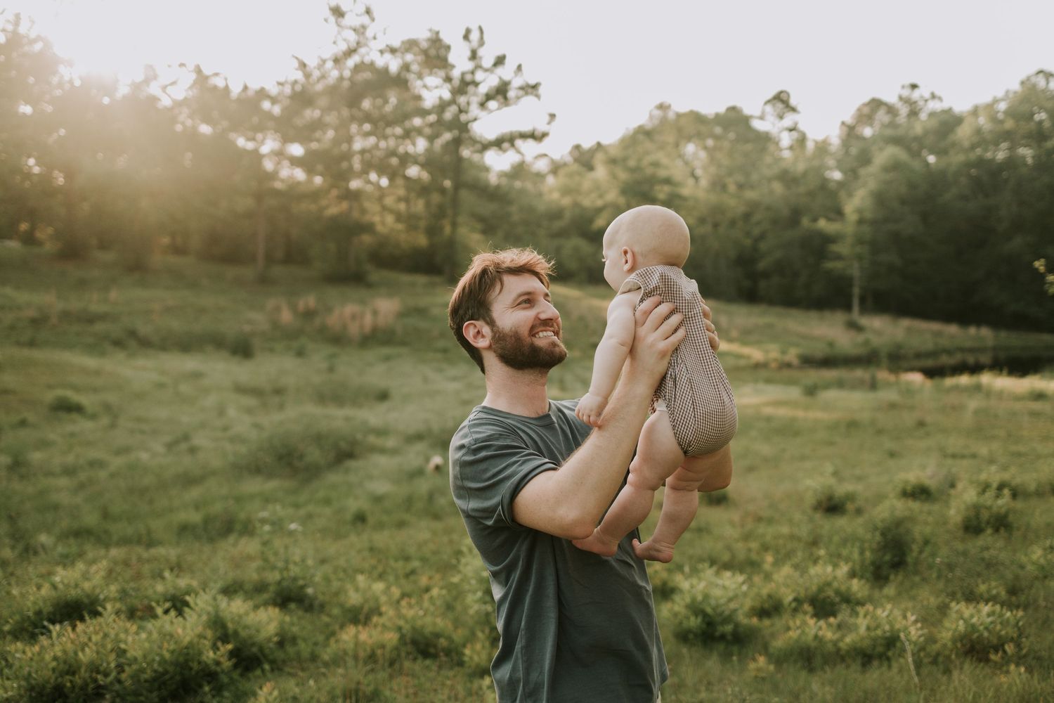 A father lifts a baby high in the air during golden hour in a grassy field.