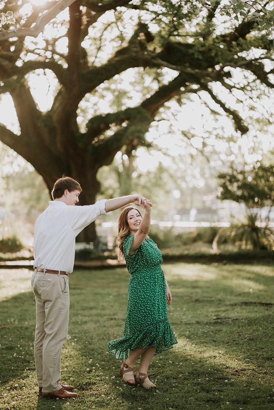 A couple dances playfully under a large oak tree during a sunny afternoon photoshoot.