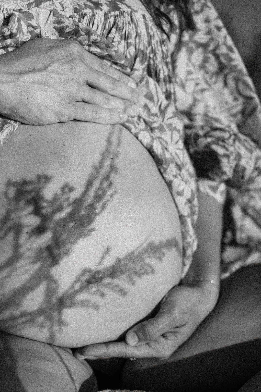 Black and white close-up of a pregnant belly with shadows cast from leaves creating a beautiful pattern.