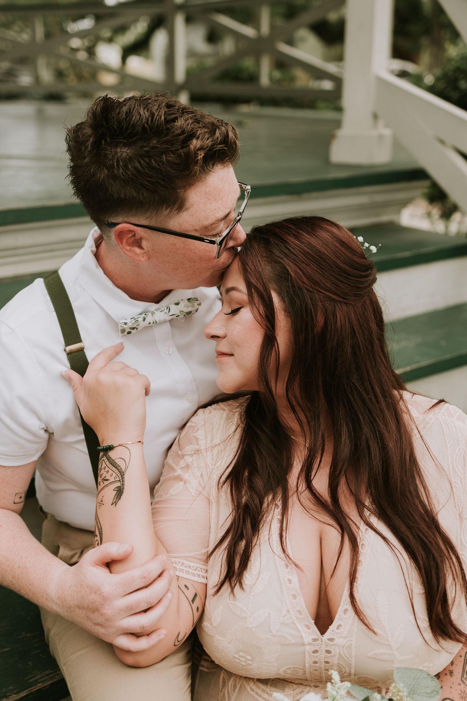 A groom in suspenders and floral bow tie kisses his bride on the forehead during a romantic outdoor wedding photo.