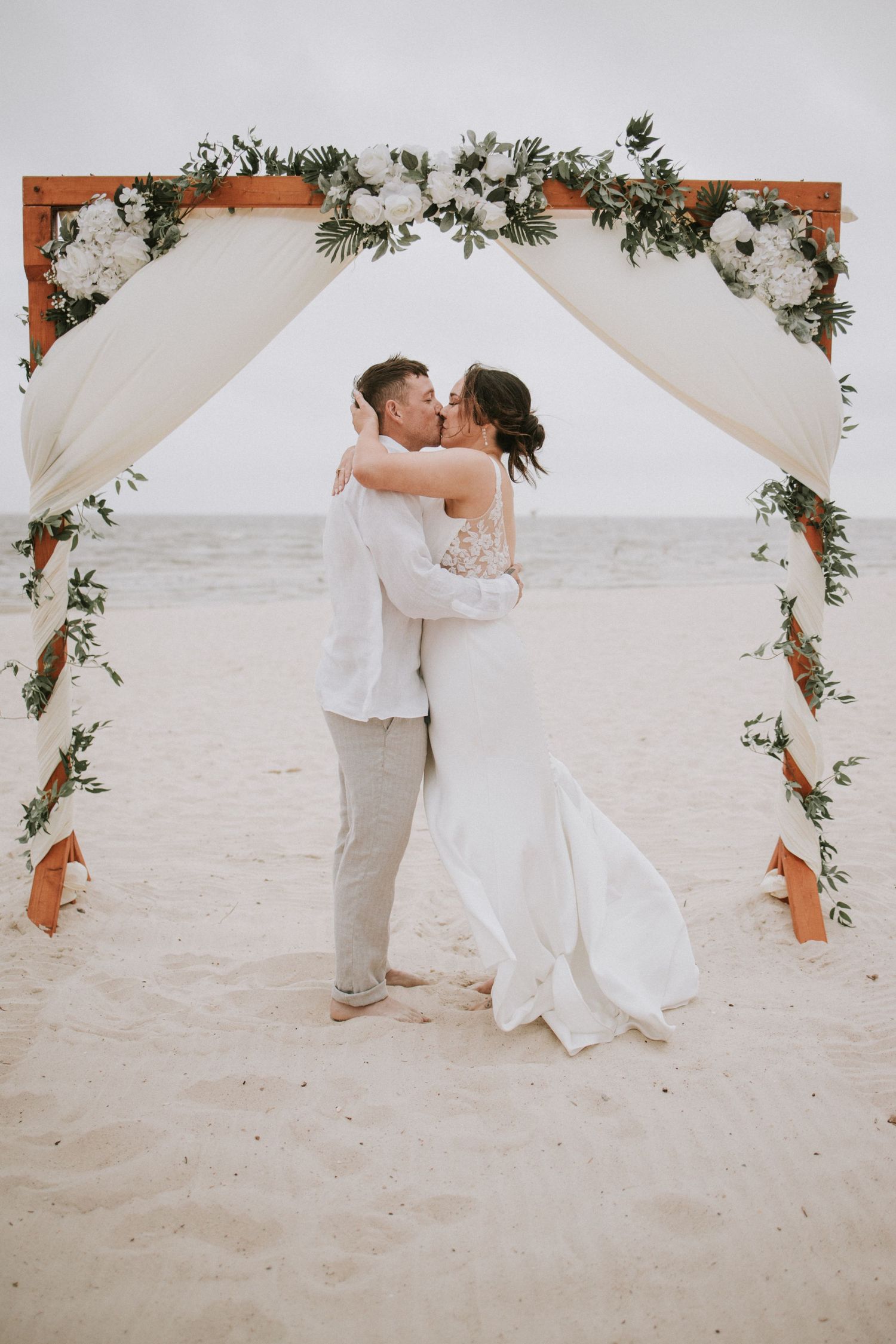 Bride and groom share first kiss under floral arch on sandy beach at their romantic outdoor wedding ceremony.