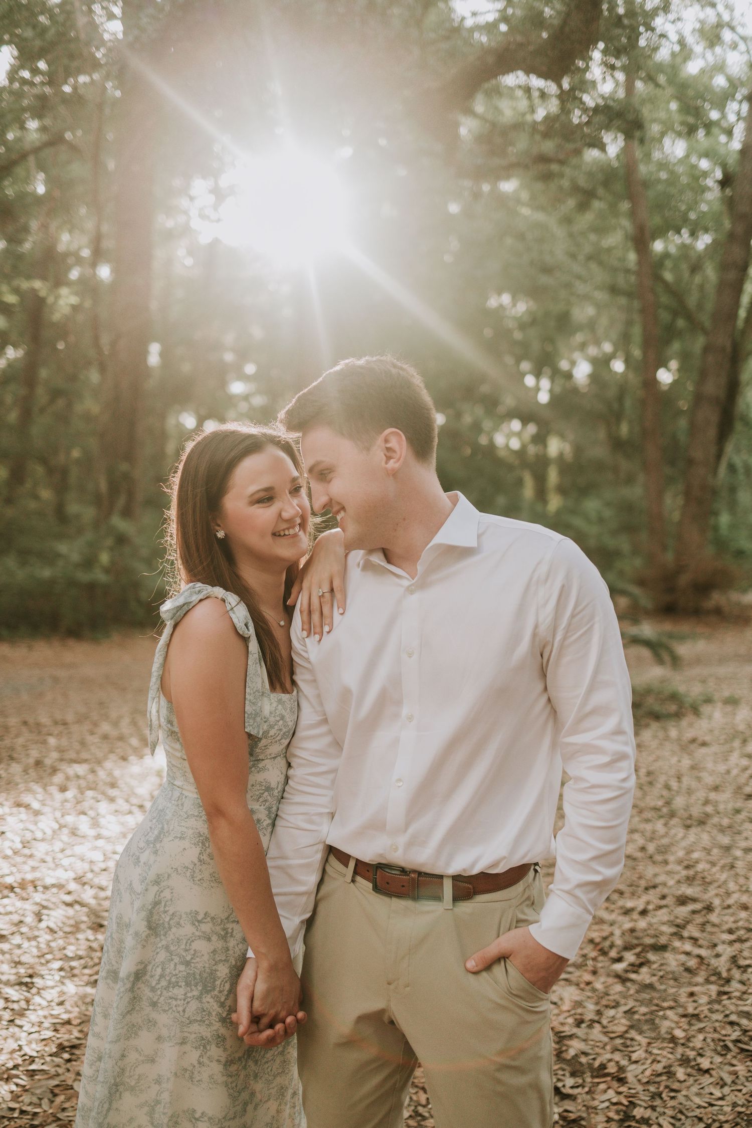Couple smiling and holding hands in a sunlit forest, woman in floral dress, man in white shirt and khaki pants.