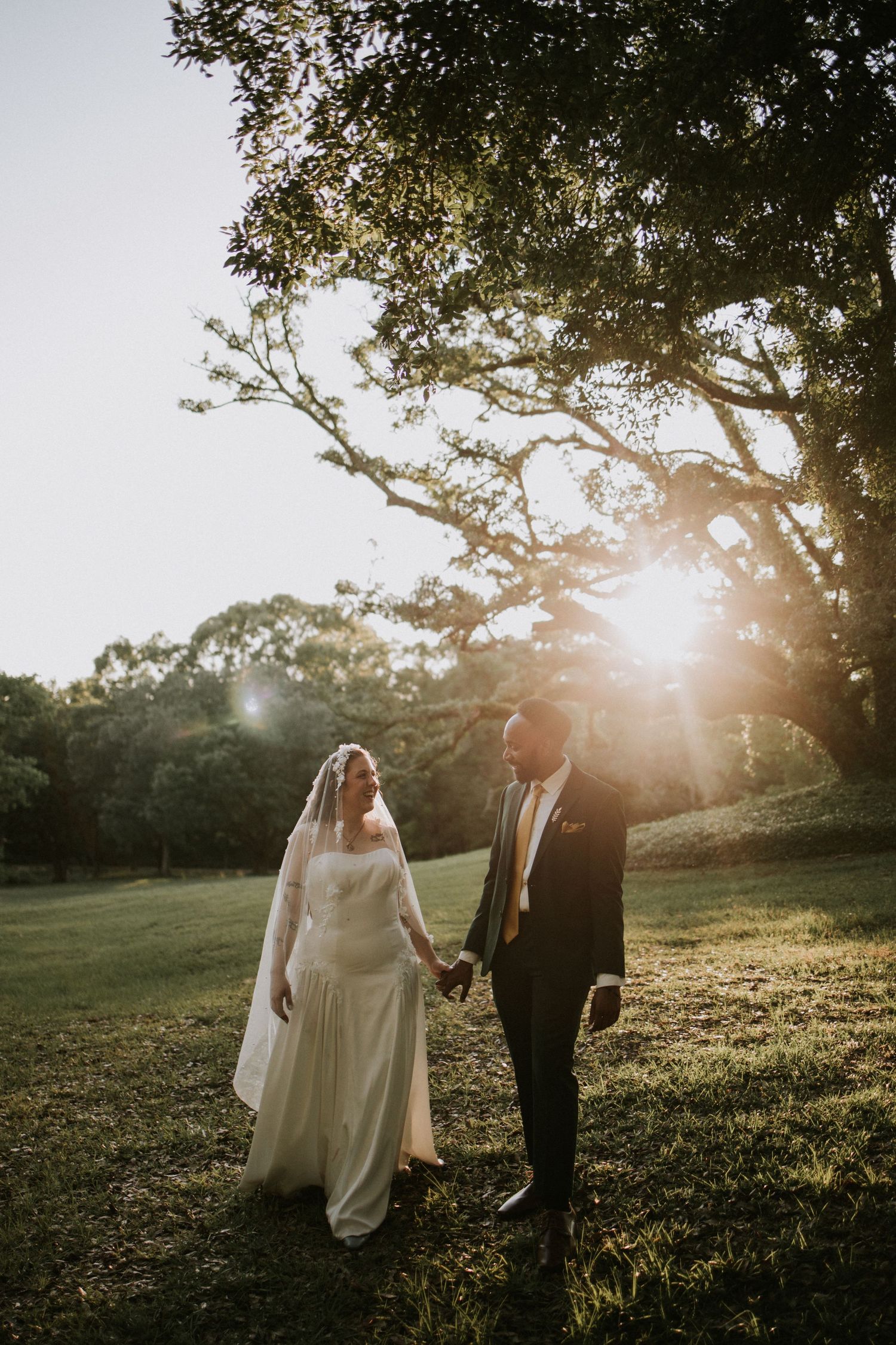 A bride and groom walk hand in hand through a sunlit park, with golden hour light streaming through lush green trees.