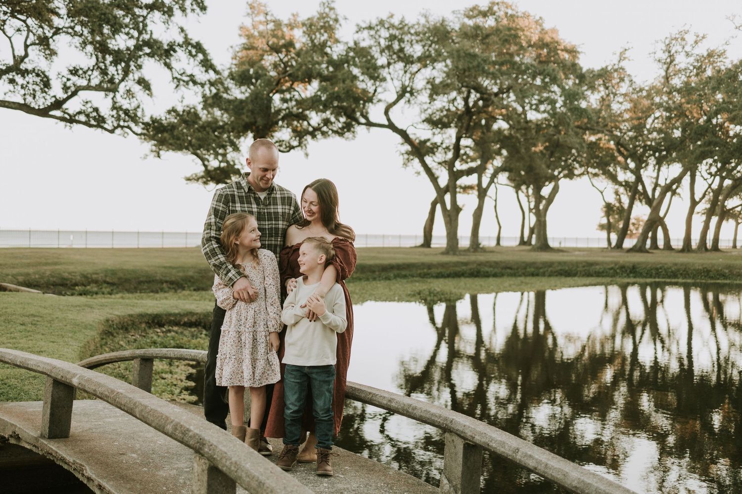 Family of five posing on a wooden bridge near a reflective pond surrounded by lush green trees at golden hour.