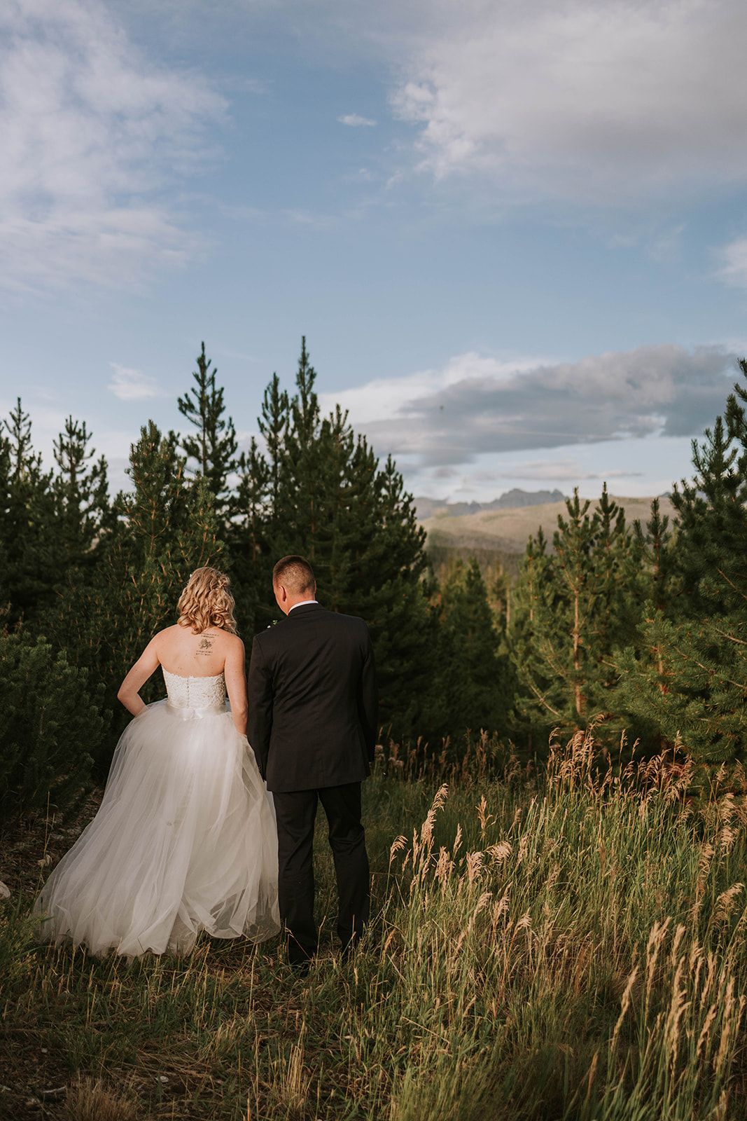 A couple in formal wedding attire stands together on a hillside surrounded by evergreen trees and tall grass at sunset.