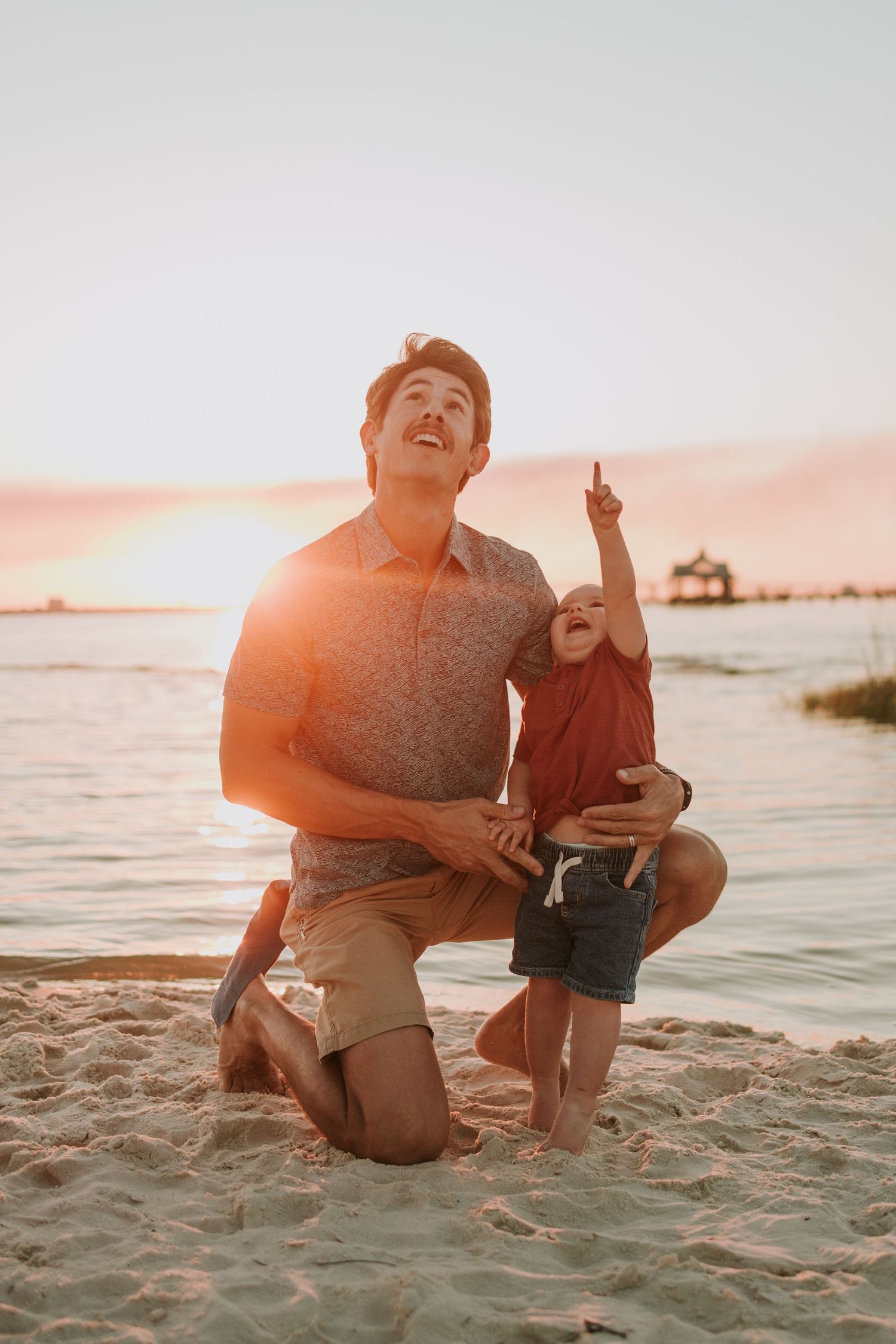 Father and young child enjoying a golden sunset on the beach, with child pointing up at the sky.