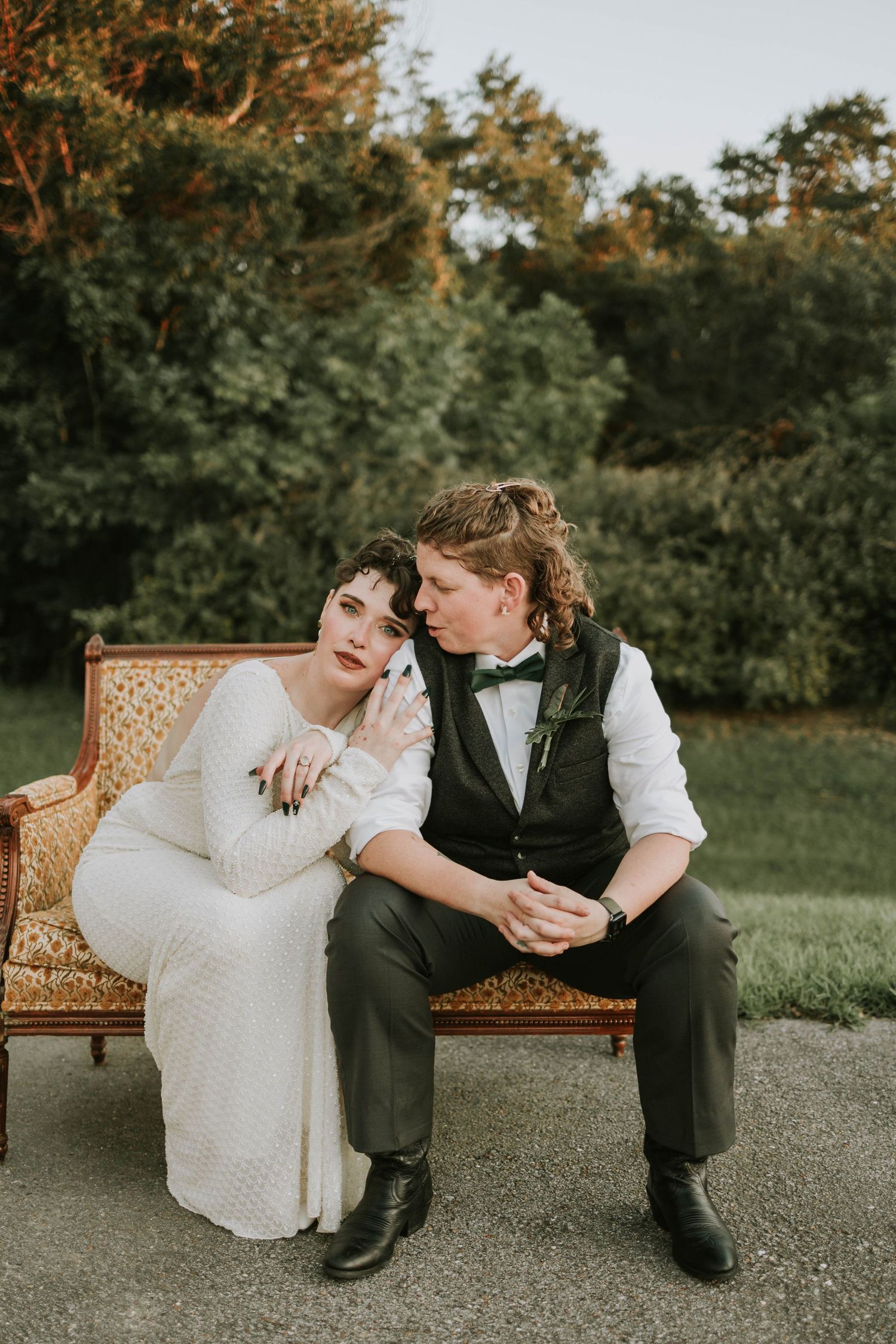 Bride and groom sitting on vintage ornate sofa outdoors, sharing an intimate moment surrounded by lush green trees.