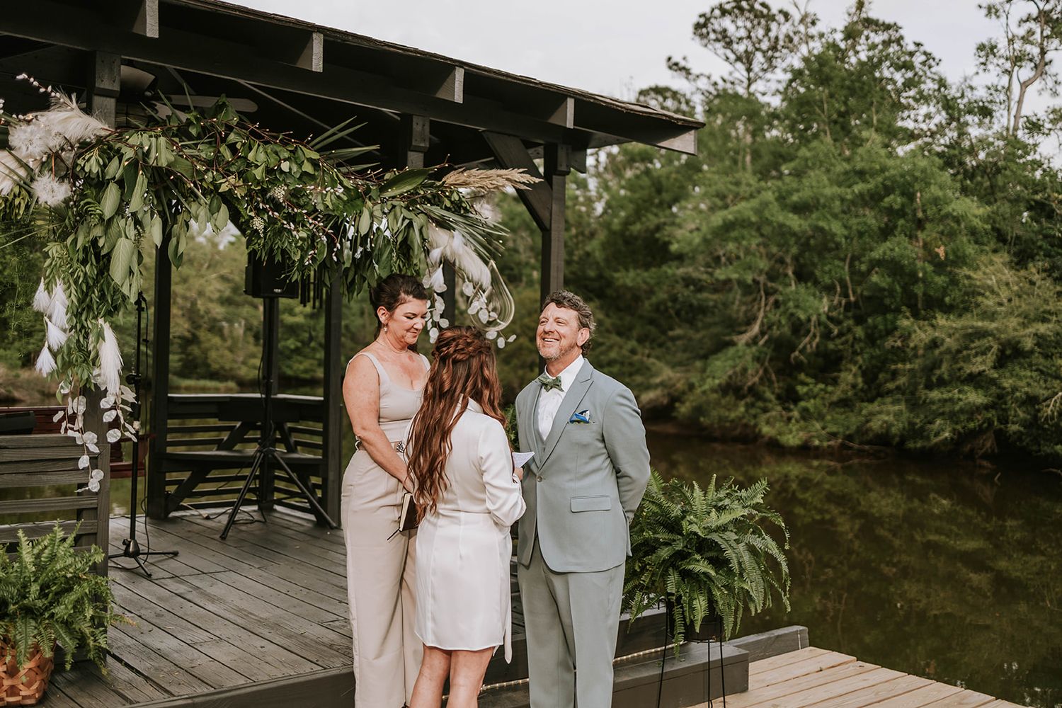 Romantic outdoor wedding ceremony at rustic wooden gazebo decorated with greenery beside tranquil pond.